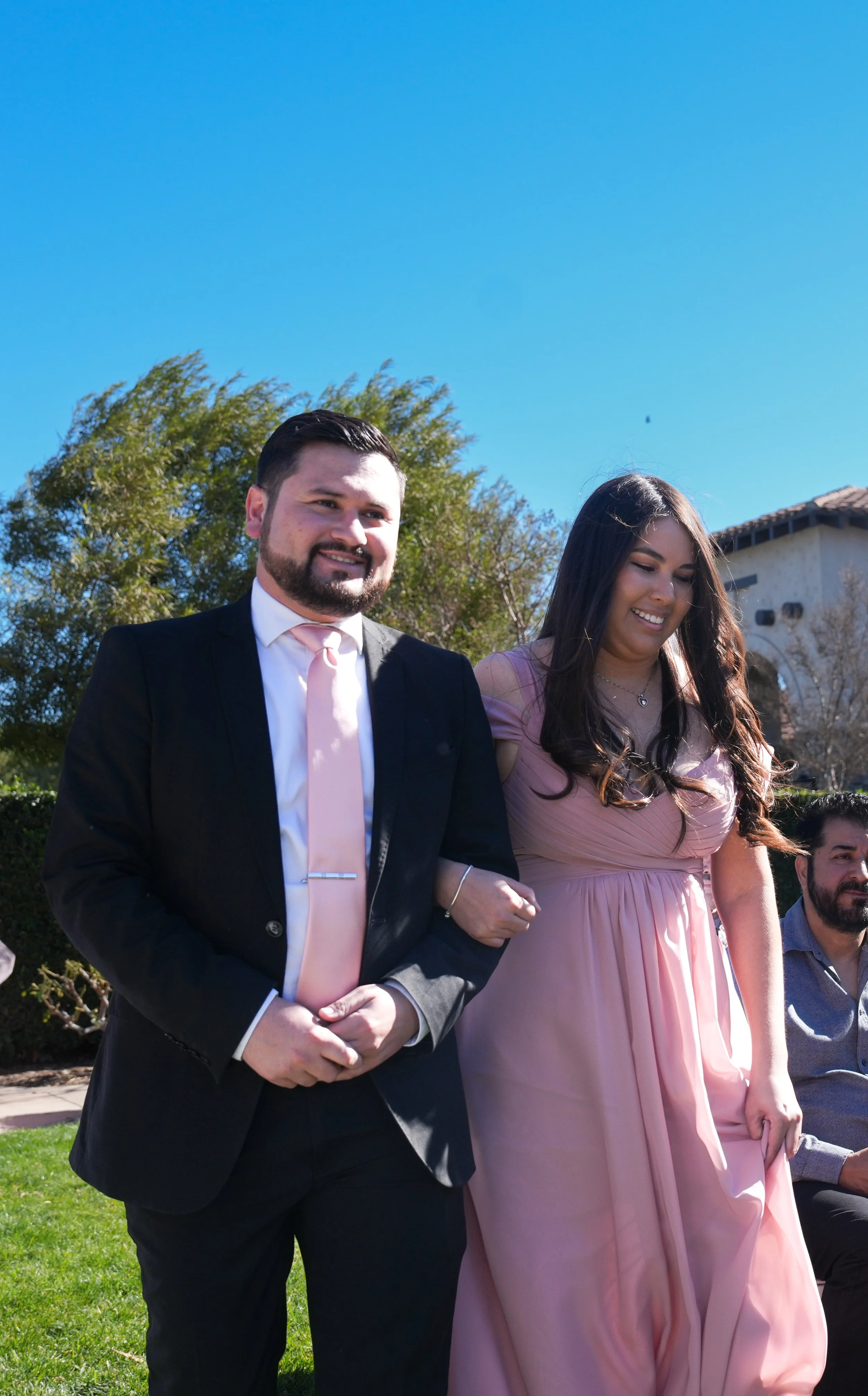 A man in a black suit with a pink tie and a woman in a pink dress participating in an outdoor event on a sunny day.