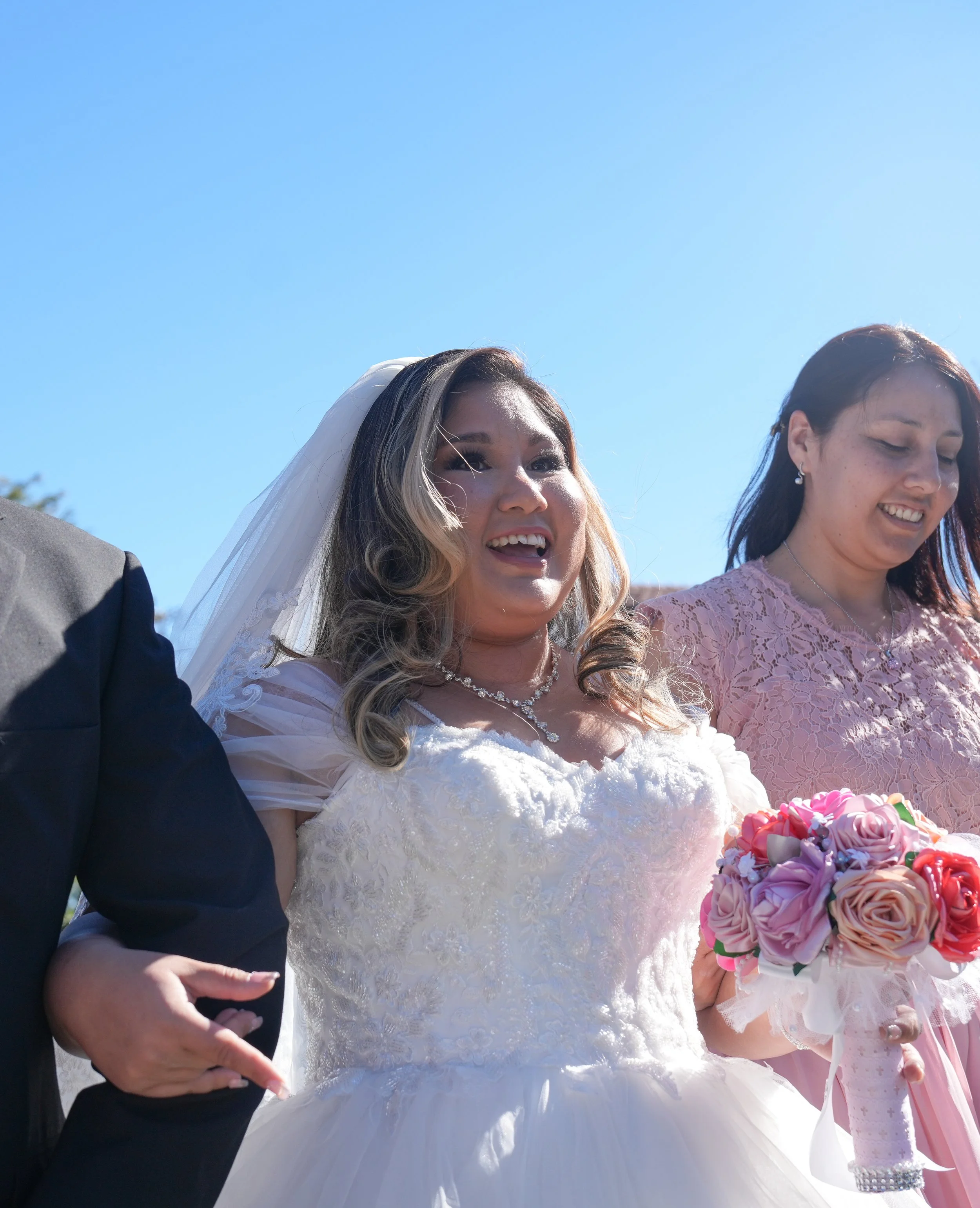 Bride in a wedding dress holding a bouquet of pink and peach roses, smiling during her wedding celebration outdoors on a sunny day.