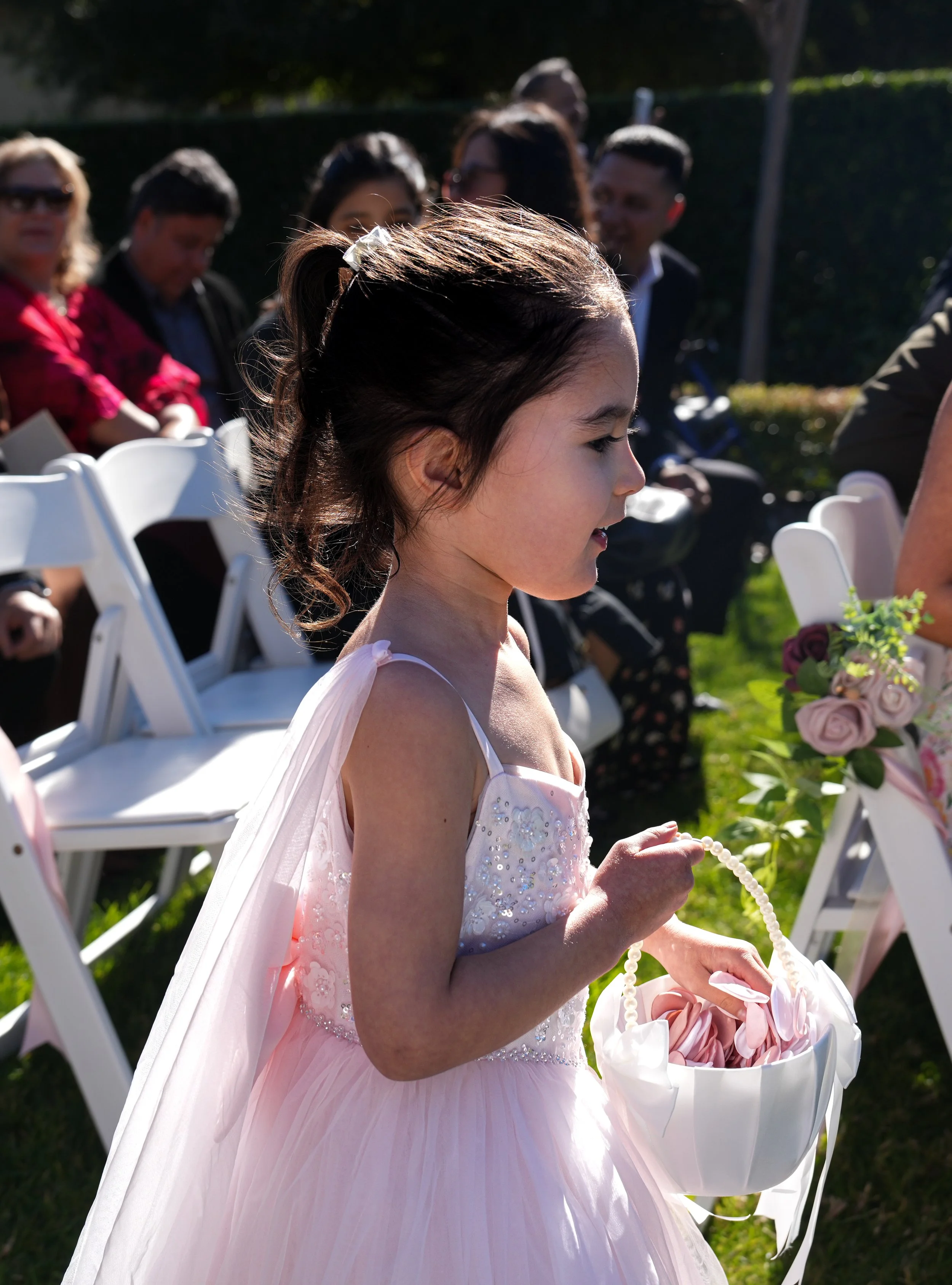 A young girl in a pink dress holding a white basket with pink fabric at an outdoor event, with seated adults in the background.