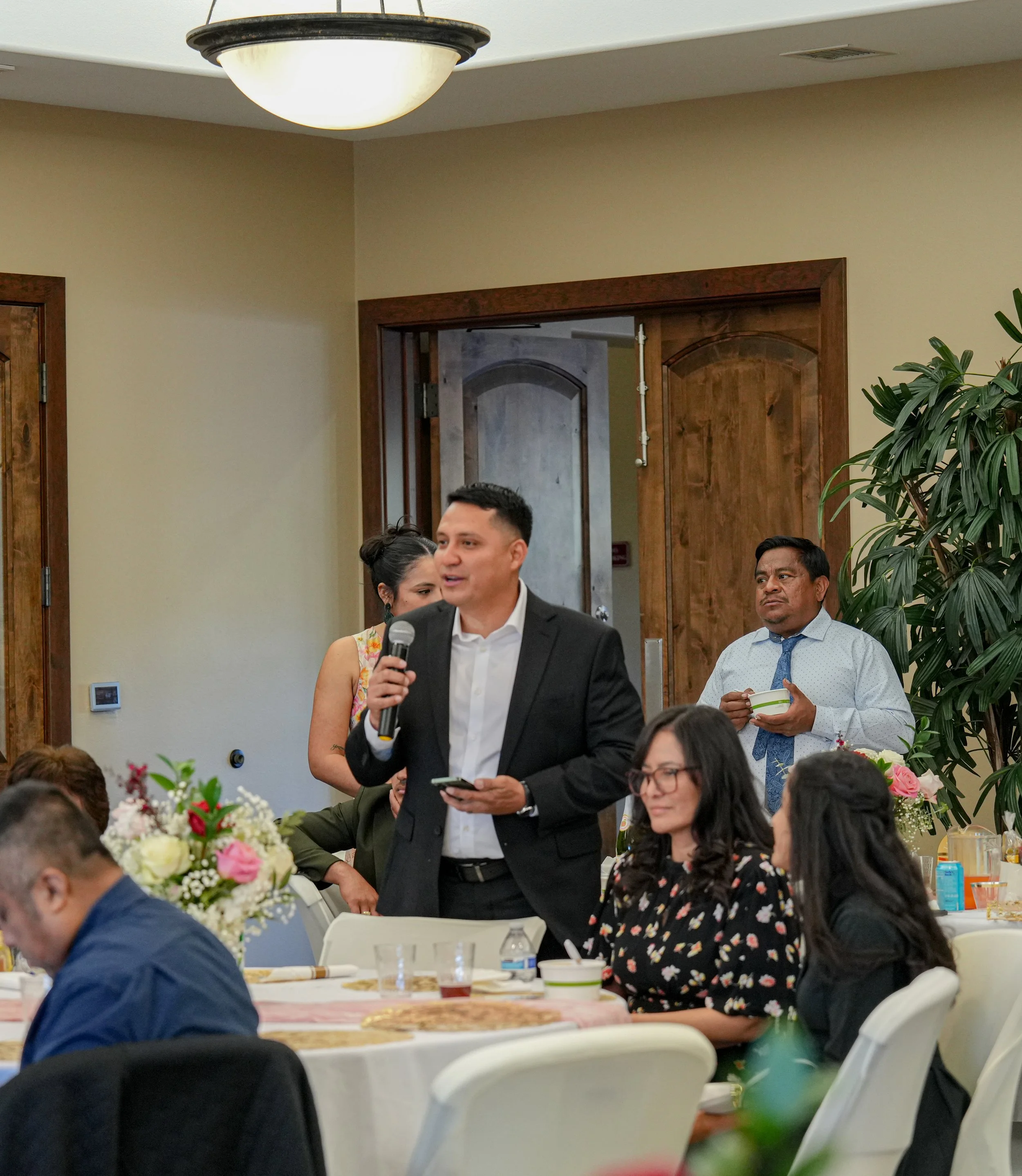A man in a black suit and white shirt speaking into a microphone at a formal event, with people seated at tables decorated with flowers in the background.