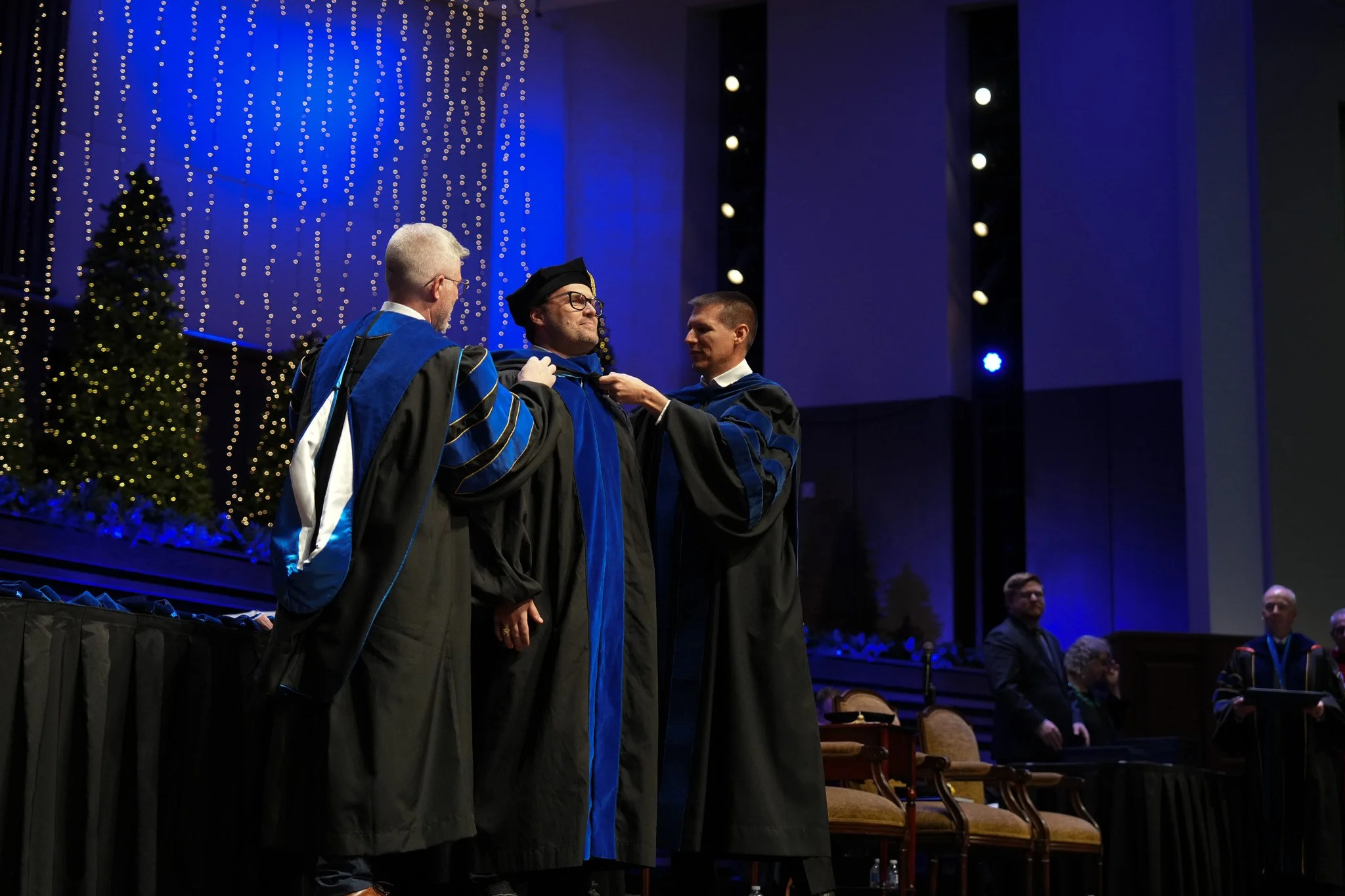 Graduation ceremony with individuals in academic regalia; one man is being awarded a stole by another man, in a decorated hall with Christmas trees and hanging lights.