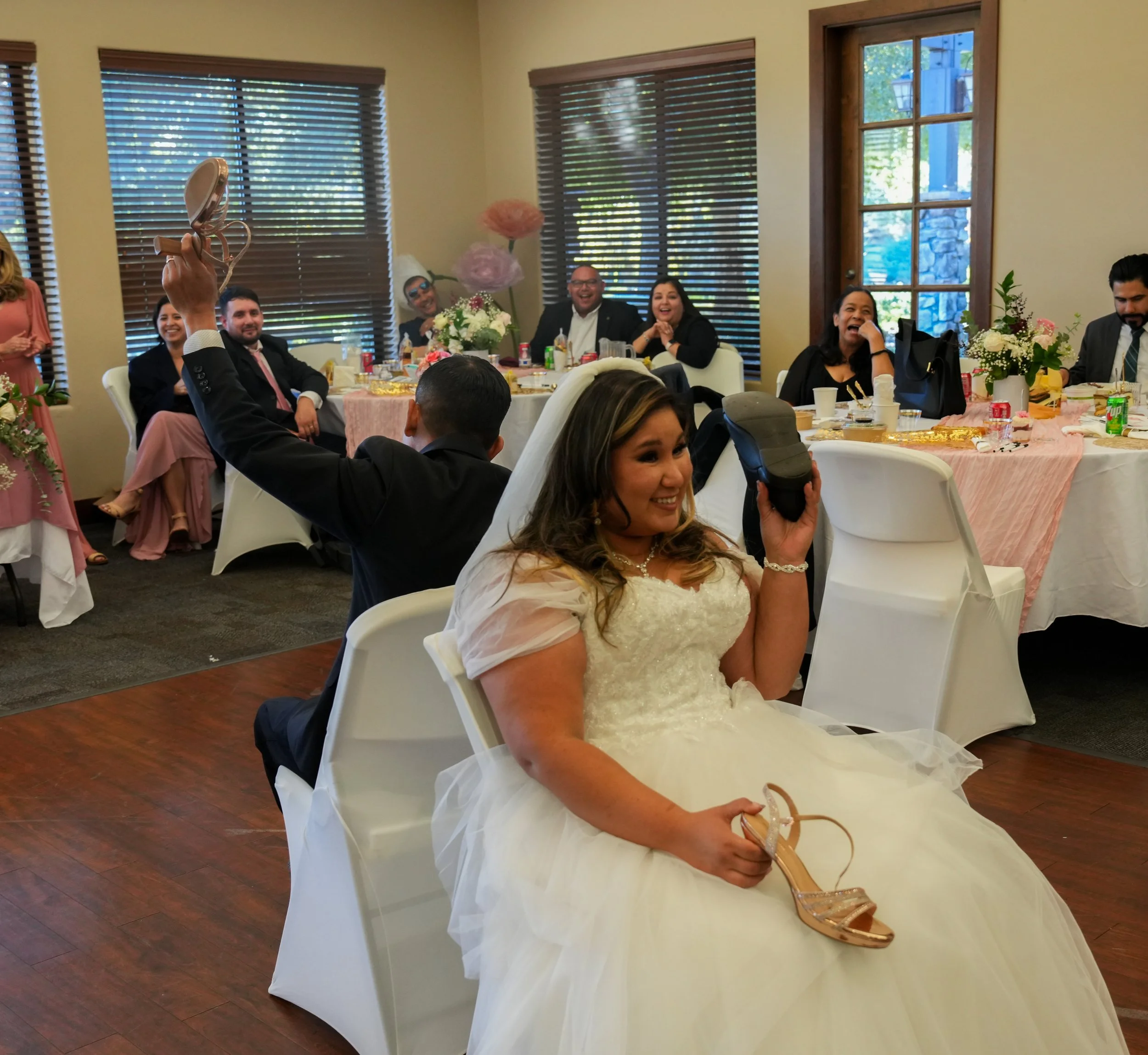 Bride in wedding dress and veil sitting on chair, smiling, holding a high heel shoe in one hand and a shoe in the other, at a wedding reception with guests seated at decorated tables, some laughing and smiling.