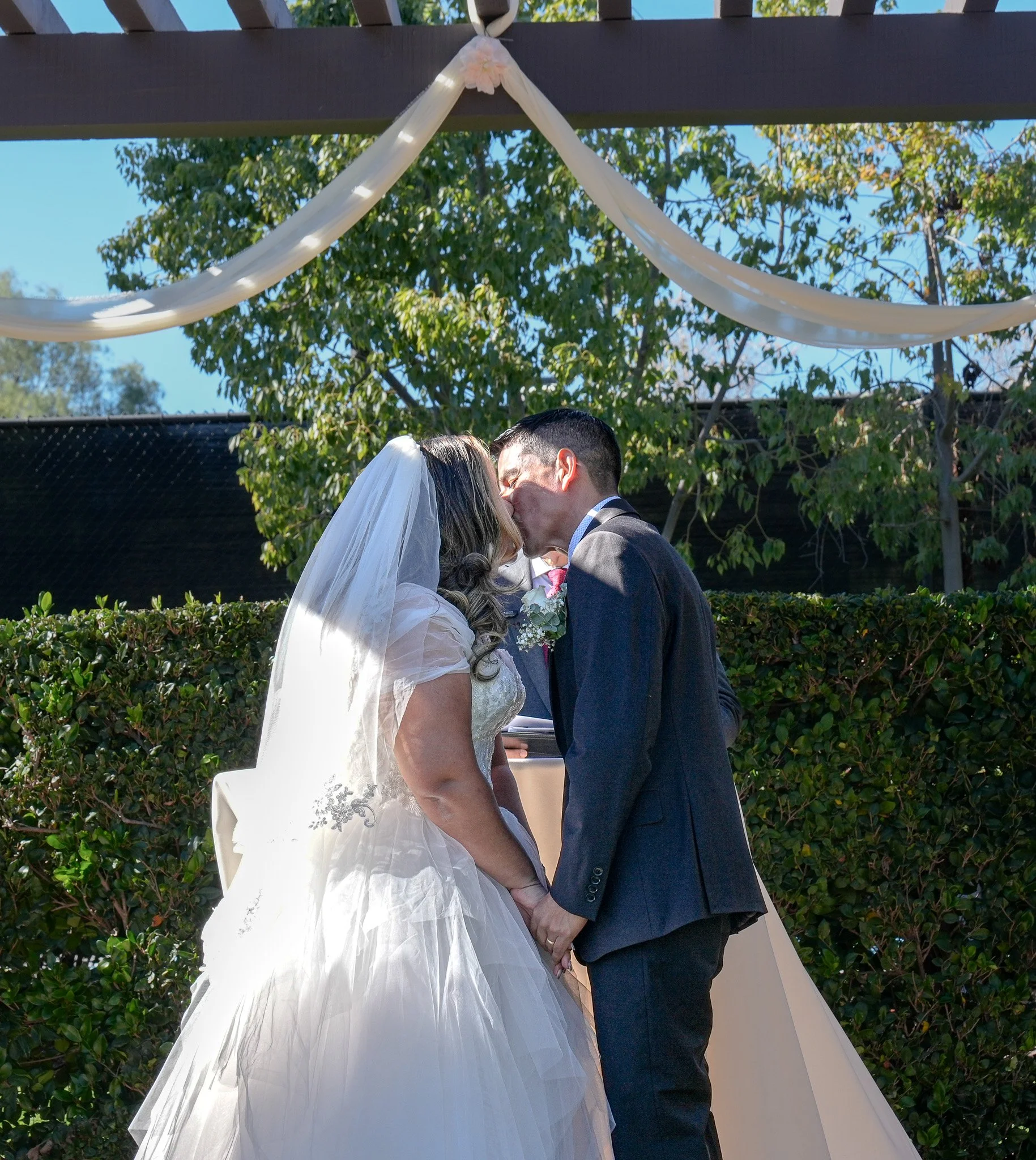 A bride and groom share a kiss during their outdoor wedding ceremony under a decorated arch.