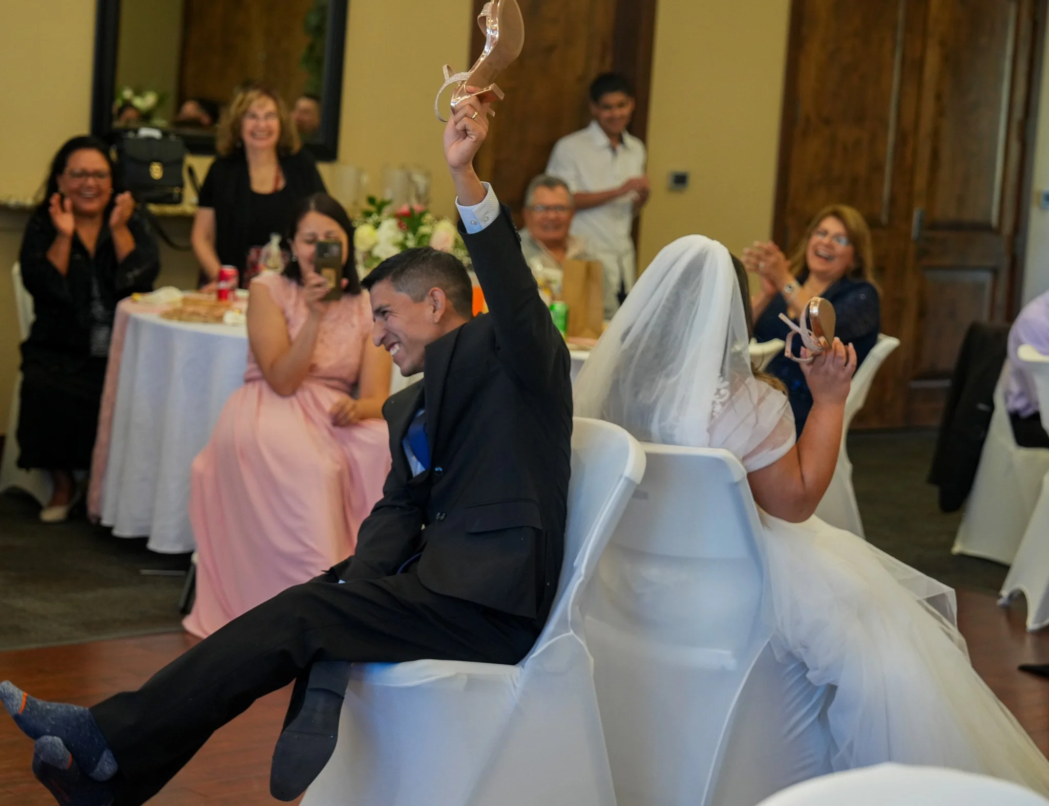 Bride and groom sitting back-to-back at a wedding reception, smiling and raising their hands with party favors, surrounded by laughing guests.