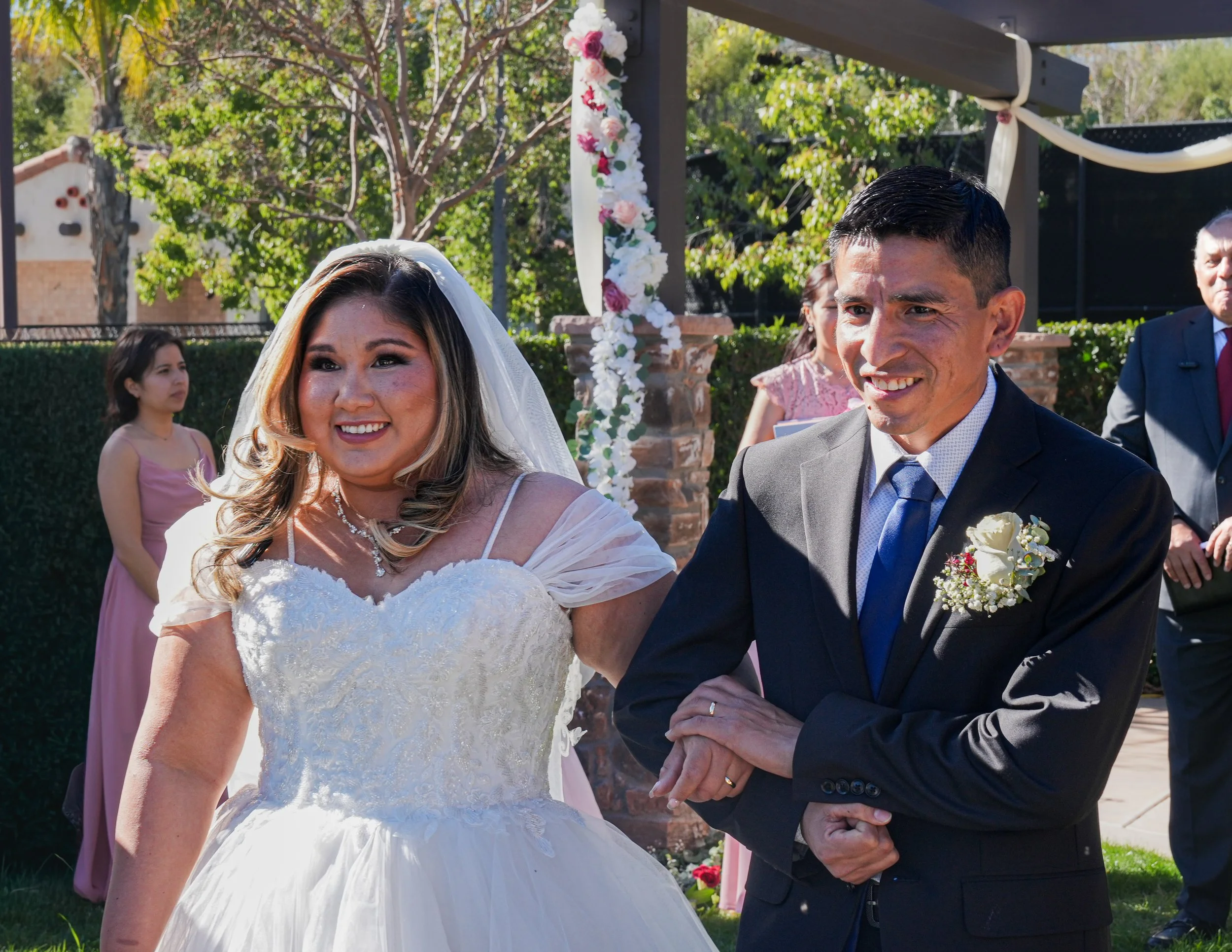 A bride and groom smiling during their wedding ceremony outdoors, with bridesmaids and groomsmen in the background.