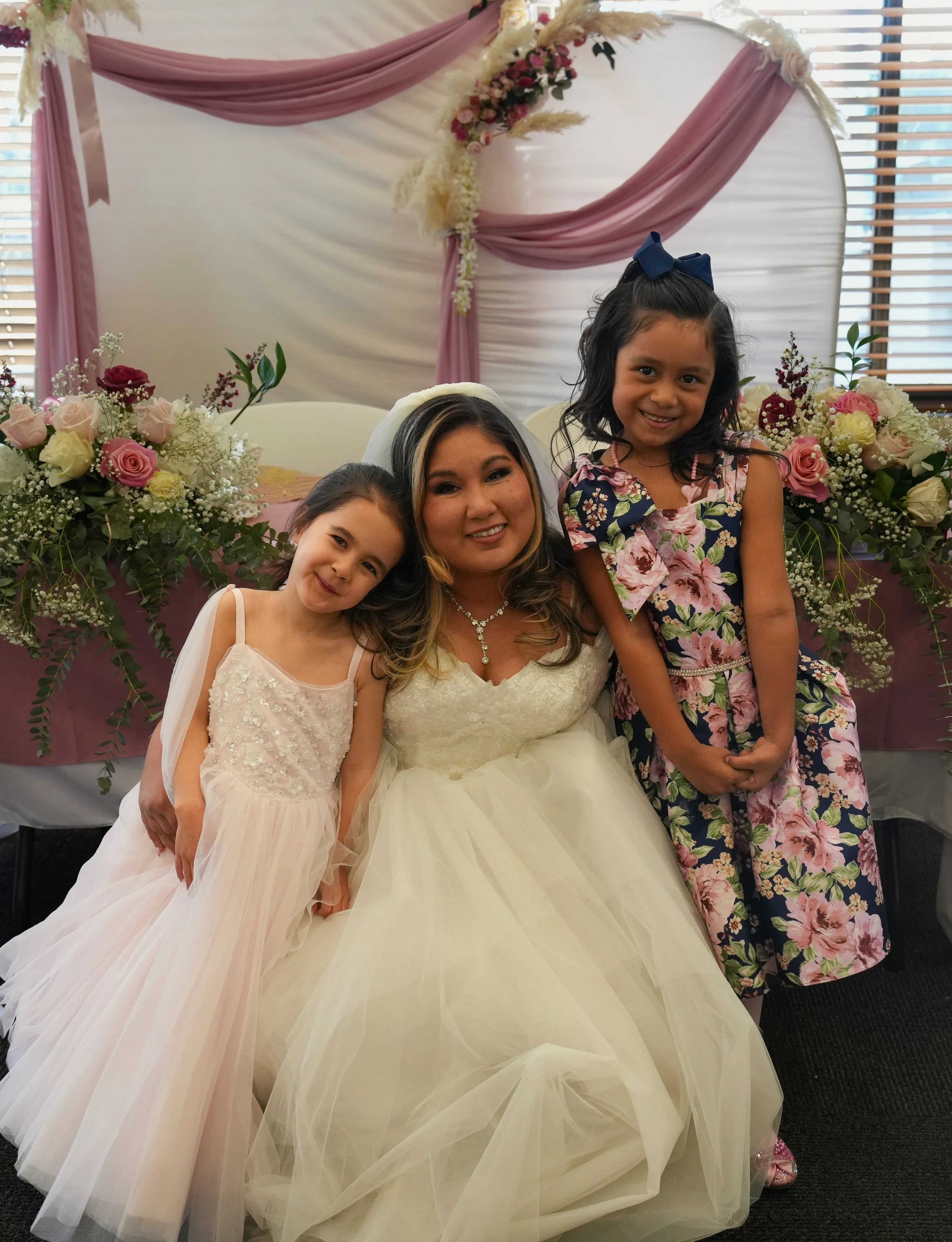 A woman in a wedding dress sitting with two young girls, all smiling at a celebration with floral decorations and pink drapery in the background.