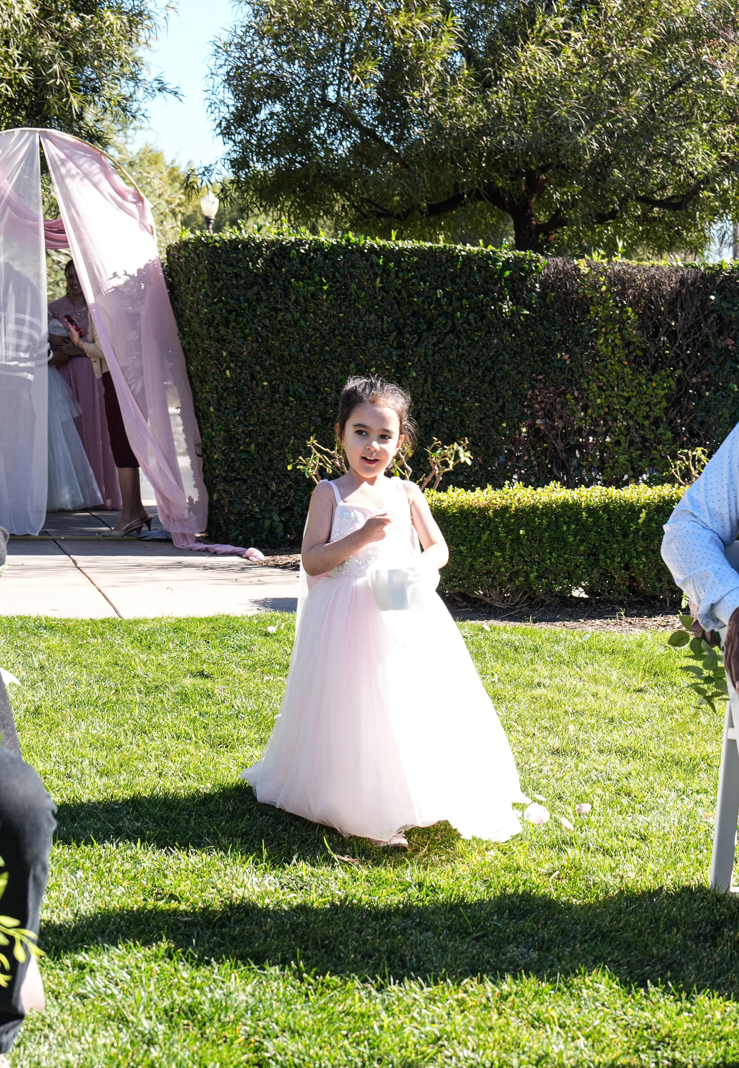 A young girl in a white dress standing outdoors on a sunny day at a wedding or formal event, with an arch and other people in the background.