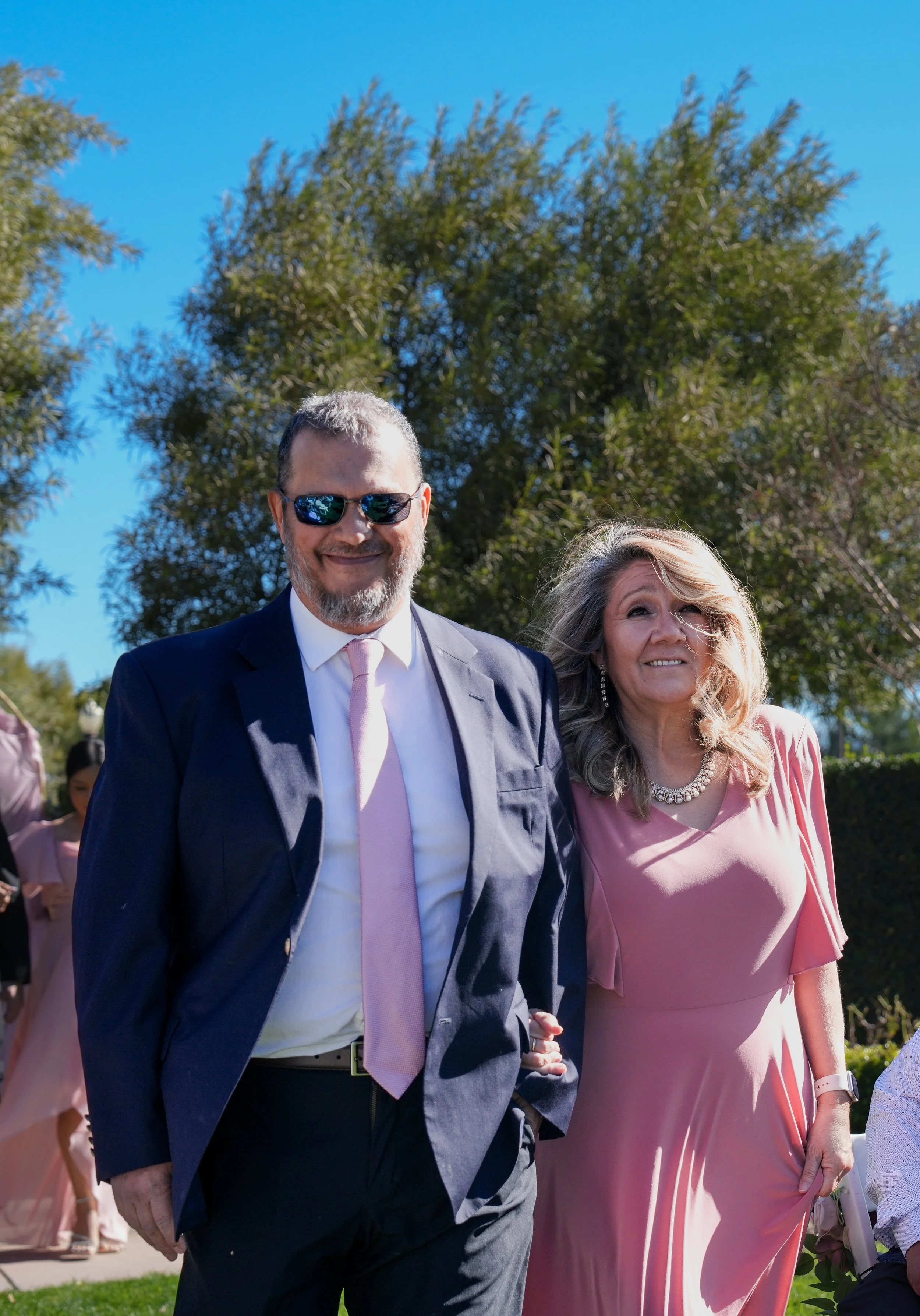 A man and woman dressed formally outdoors on a sunny day, with trees and a clear blue sky in the background. The man is wearing a dark suit, white shirt, pink tie, and sunglasses. The woman is in a pink dress with a pearl necklace and has long, wavy 