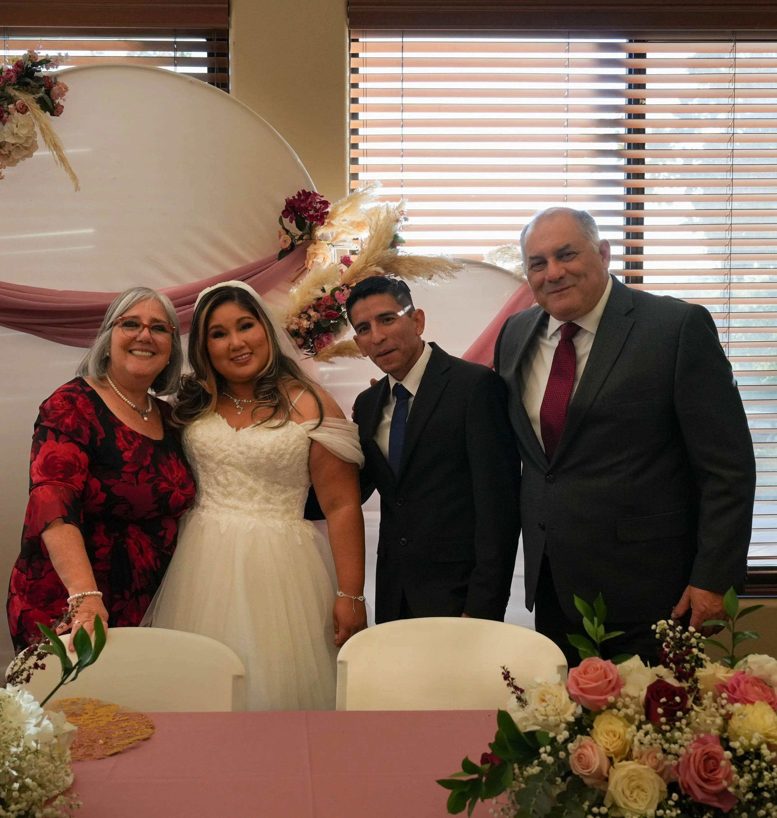 Four people standing together at a celebration, with a decorated backdrop of pink and white flowers, pampas grass, and draped fabric, in front of a window with blinds.