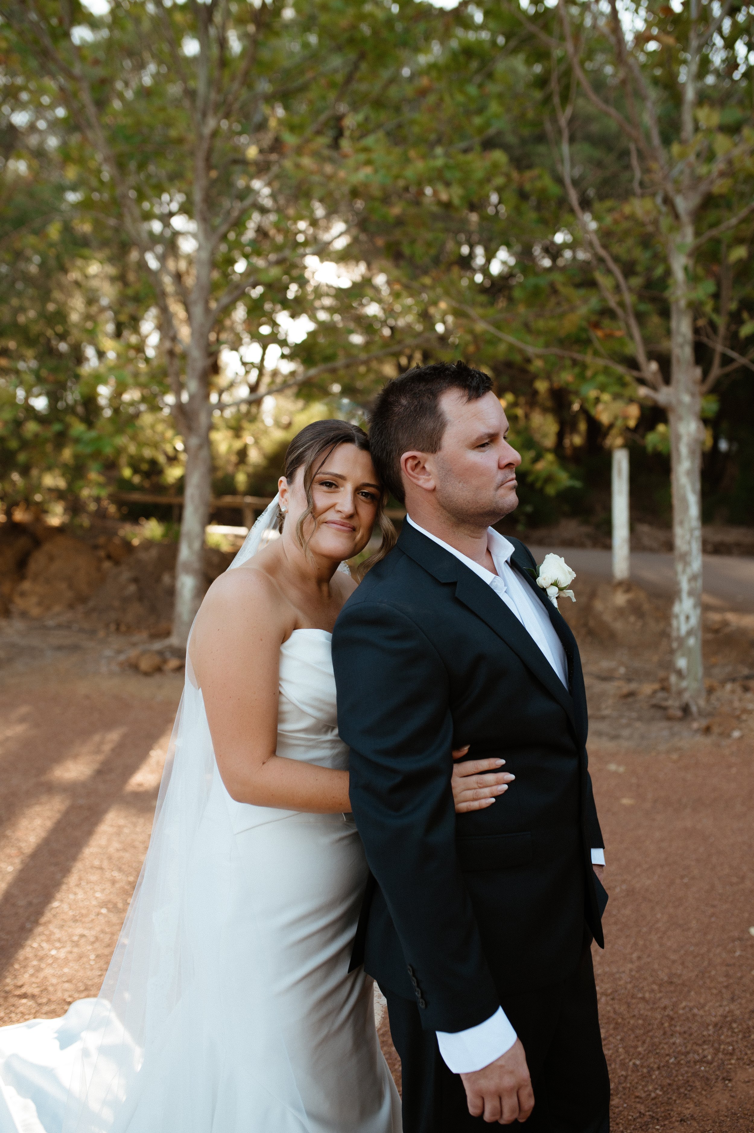 A bride and groom standing outdoors, with trees and a dirt path in the background, during their wedding celebration.