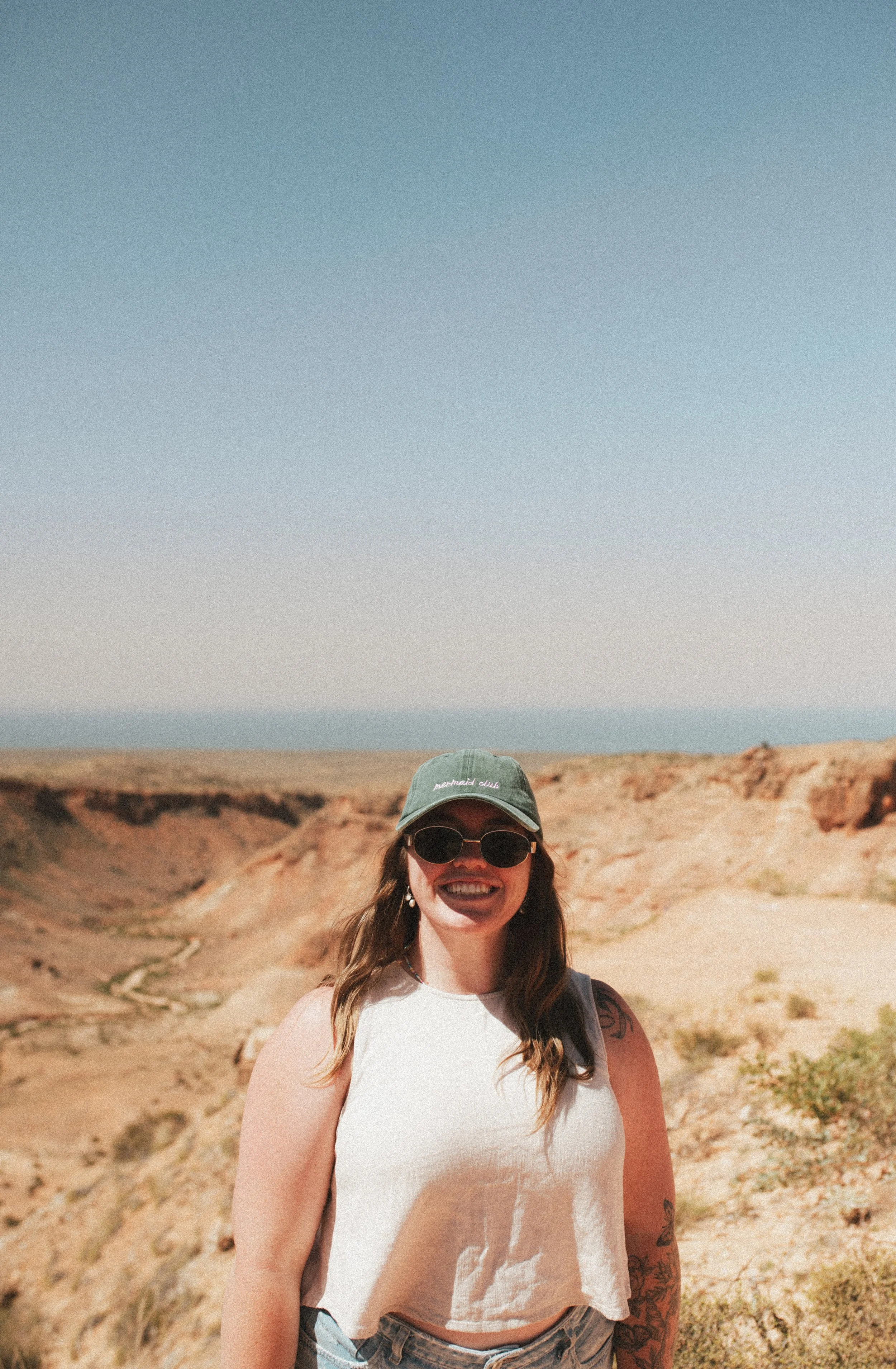 Woman smiling outdoors in desert landscape, wearing sunglasses, a cap, and a sleeveless top.