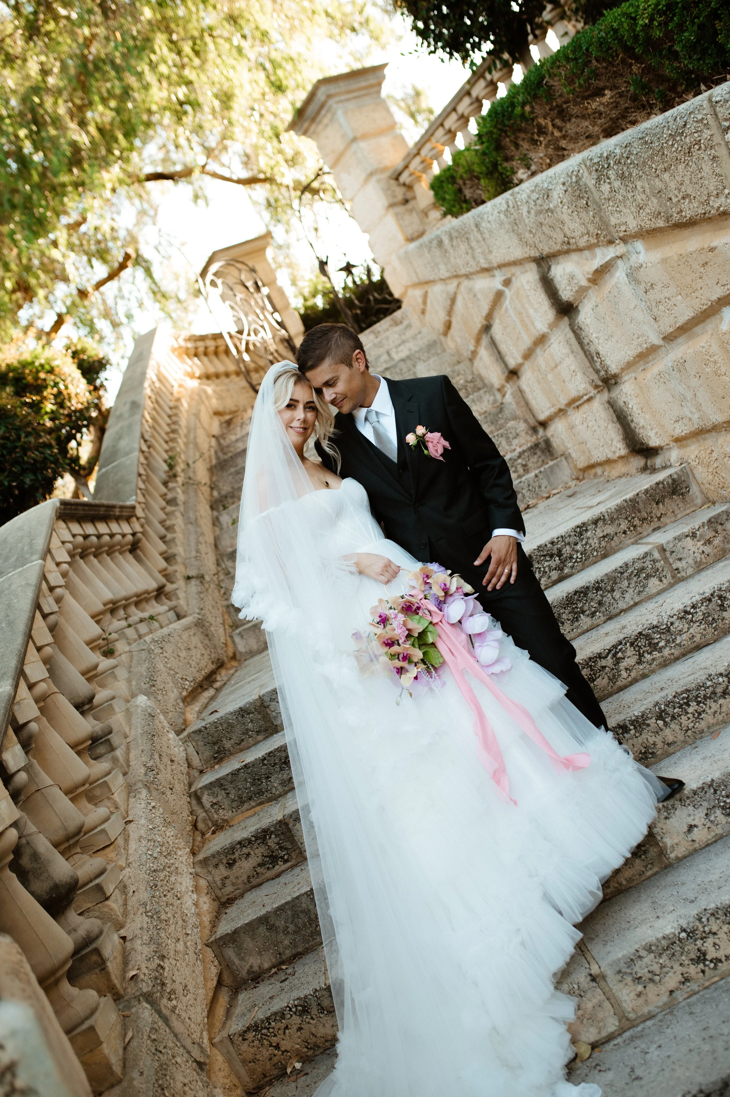 A bride and groom on stone stairs outside, with trees and historic architecture in the background. The bride is holding a bouquet of pink and purple flowers, and they are sharing a tender moment.