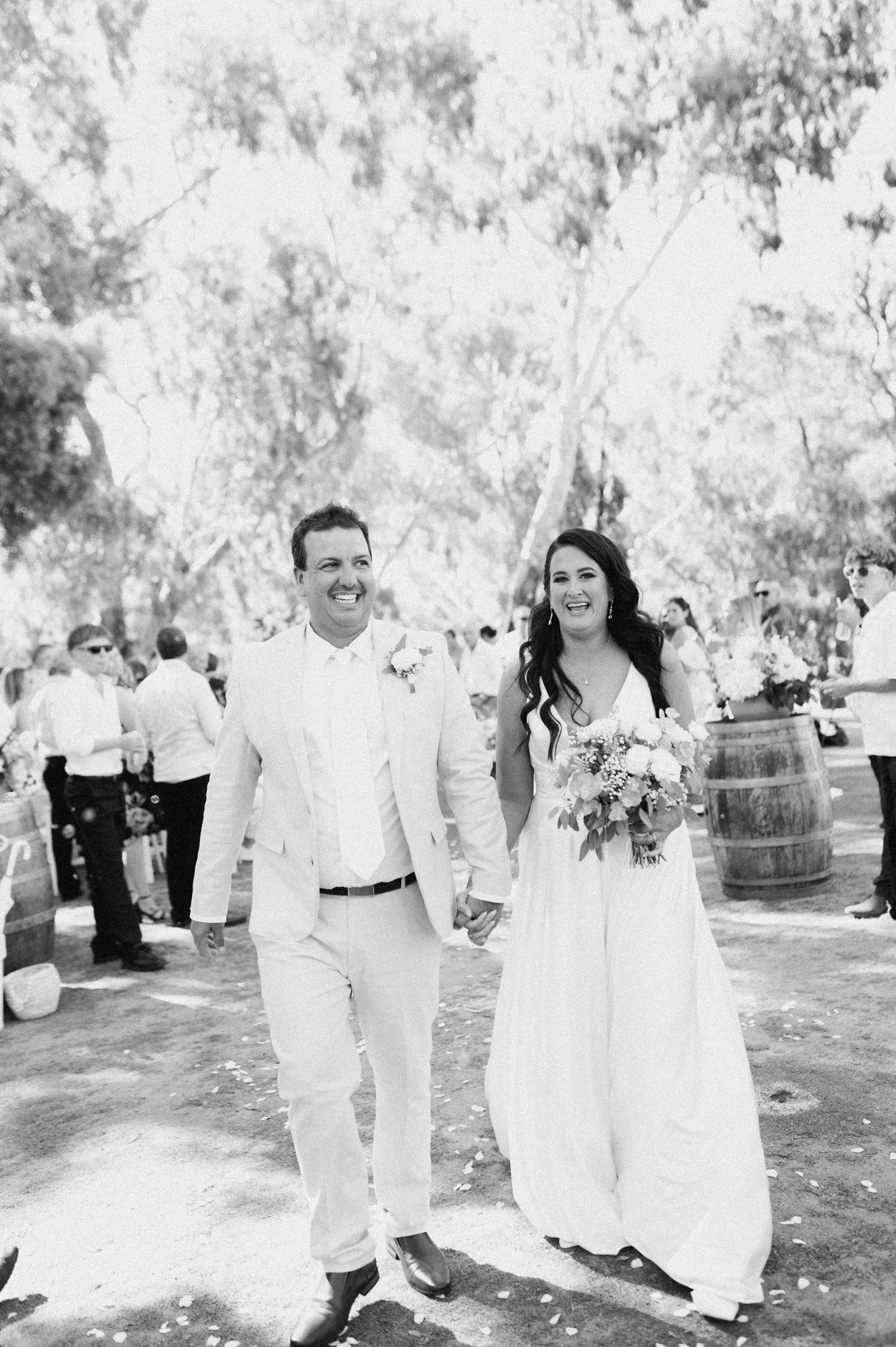 Black and white photo of a happy couple walking hand in hand at their wedding outdoors, surrounded by guests, trees, and decorative barrels.