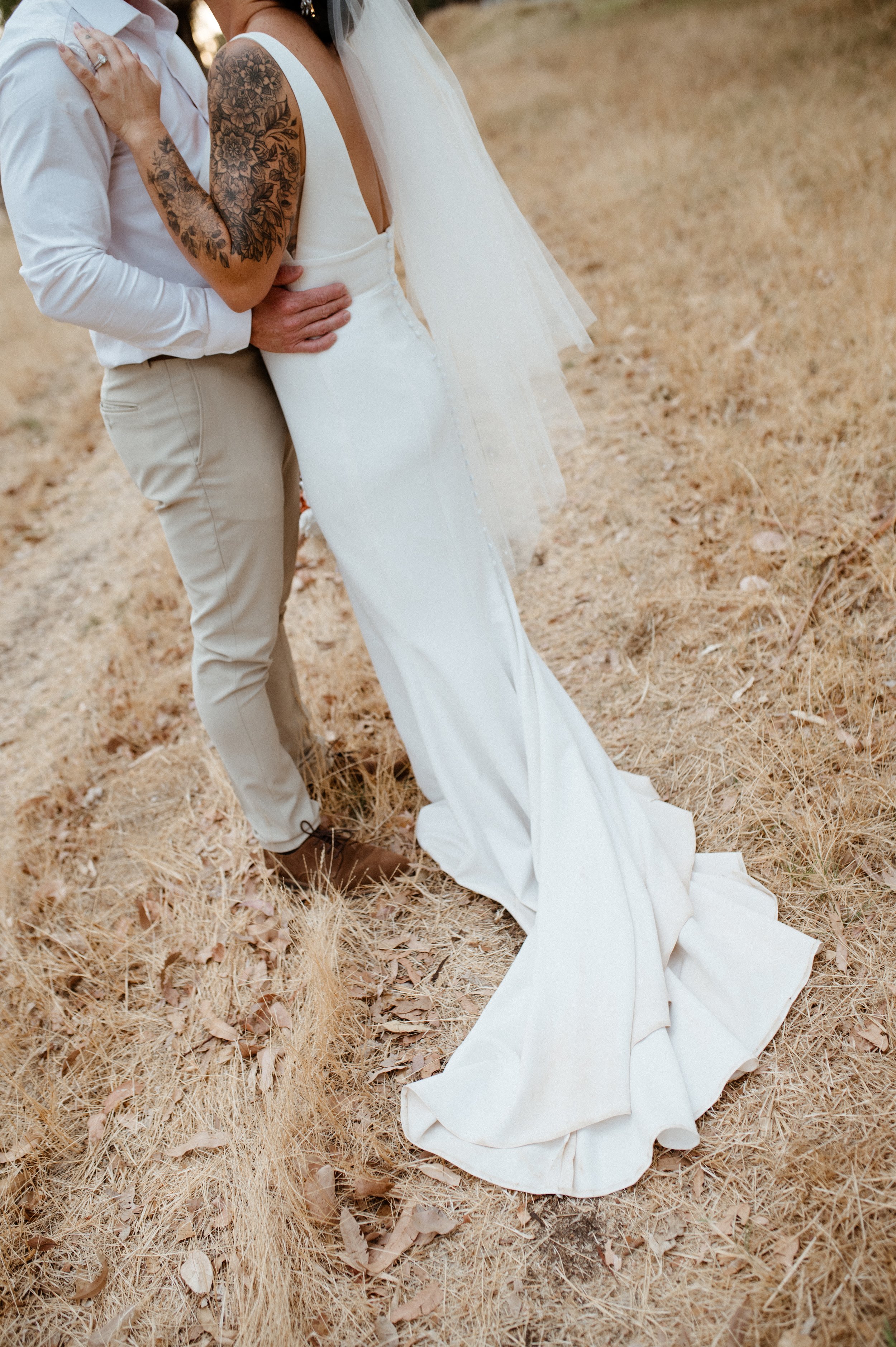 Close-up of a bride and groom standing outdoors on a dry grassy path. The bride wears a white bridal gown with a tulle veil, and the groom is dressed in a white shirt and light-colored pants. The bride has tattoos on her arms, and they are holding ea