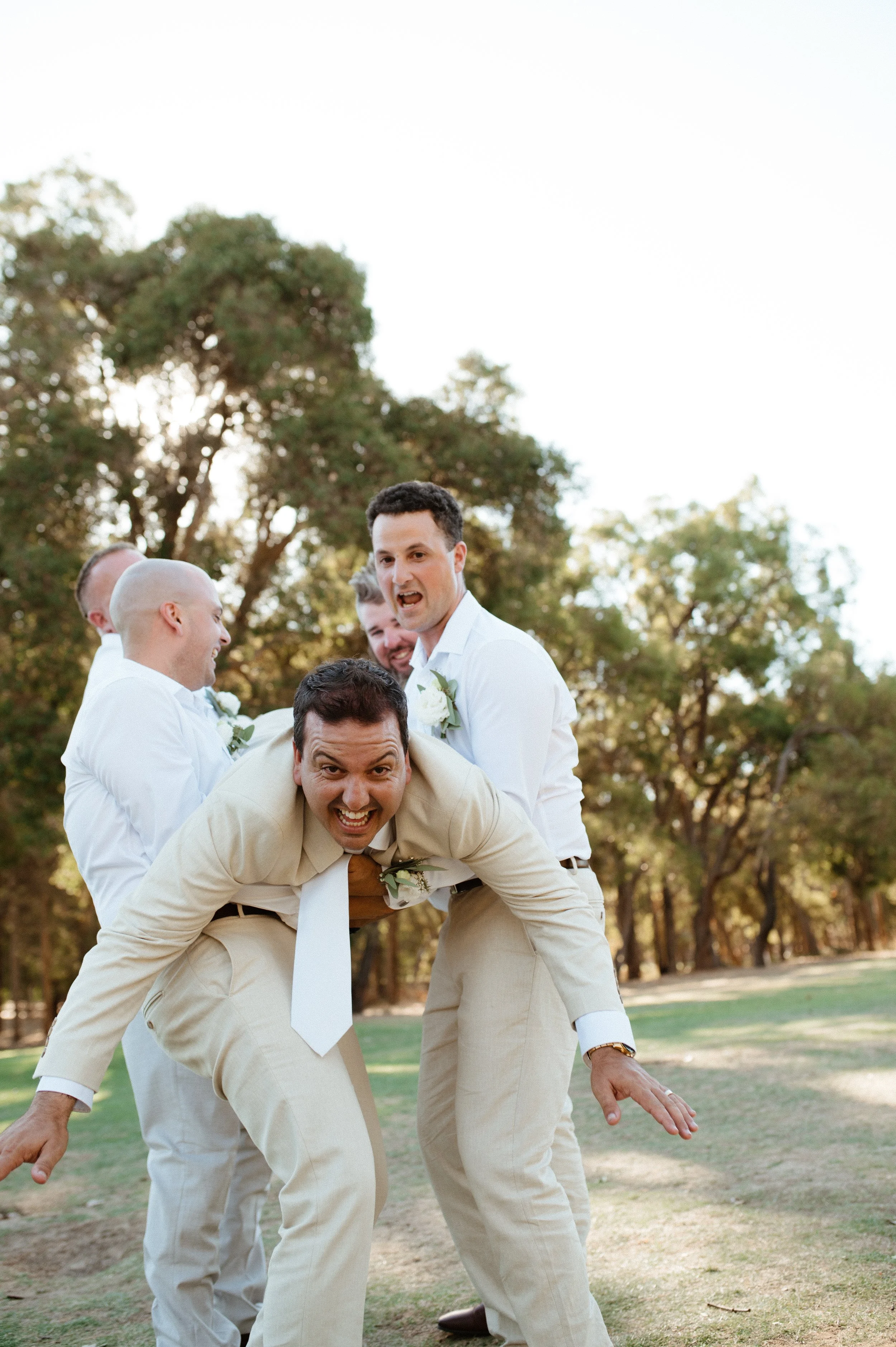 A group of men dressed in white and beige suits posing playfully outdoors with trees in the background.