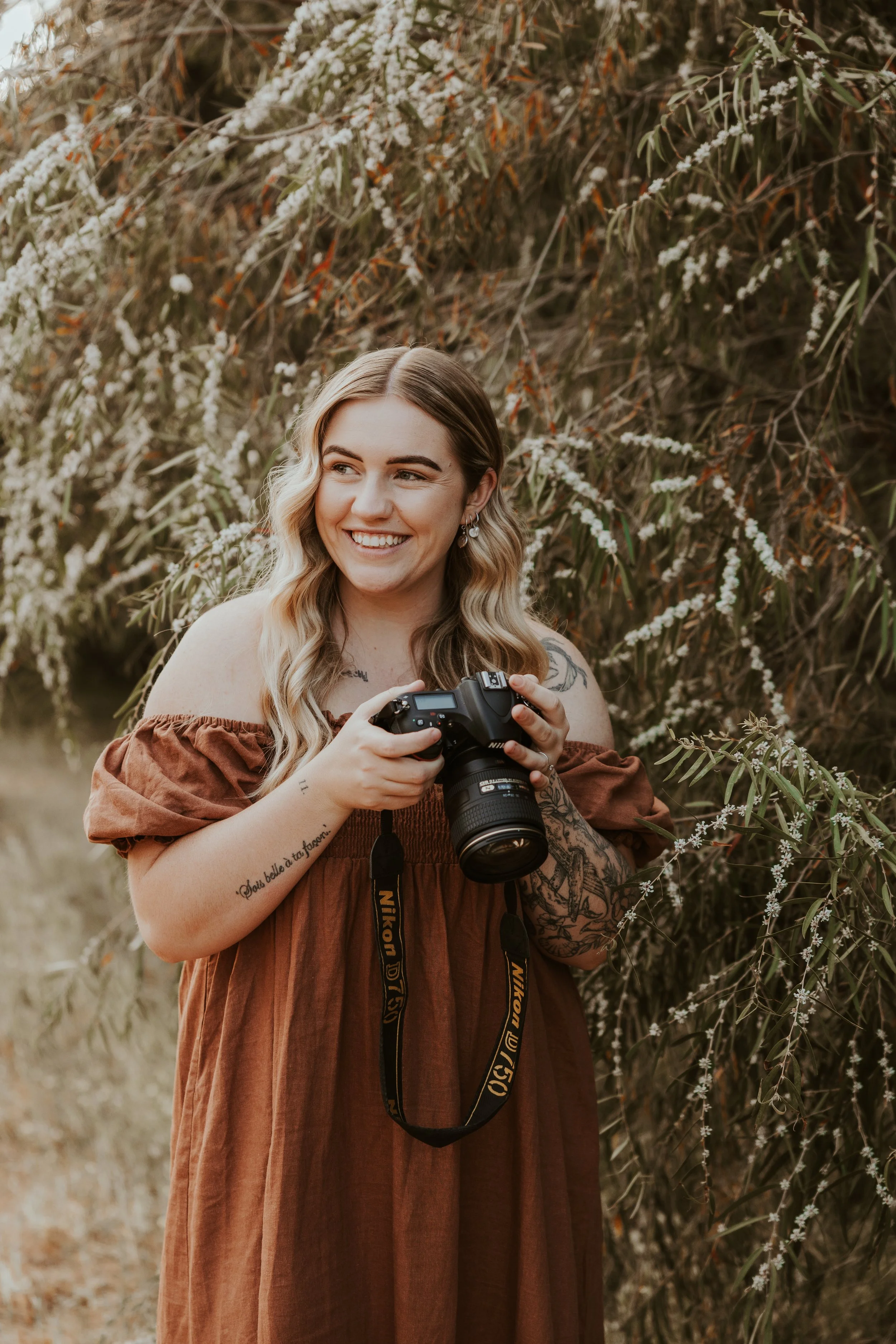 A young woman with wavy blonde hair, wearing a brown off-shoulder dress, holding a Nikon camera, standing outdoors near flowering bushes, smiling and looking to the side.