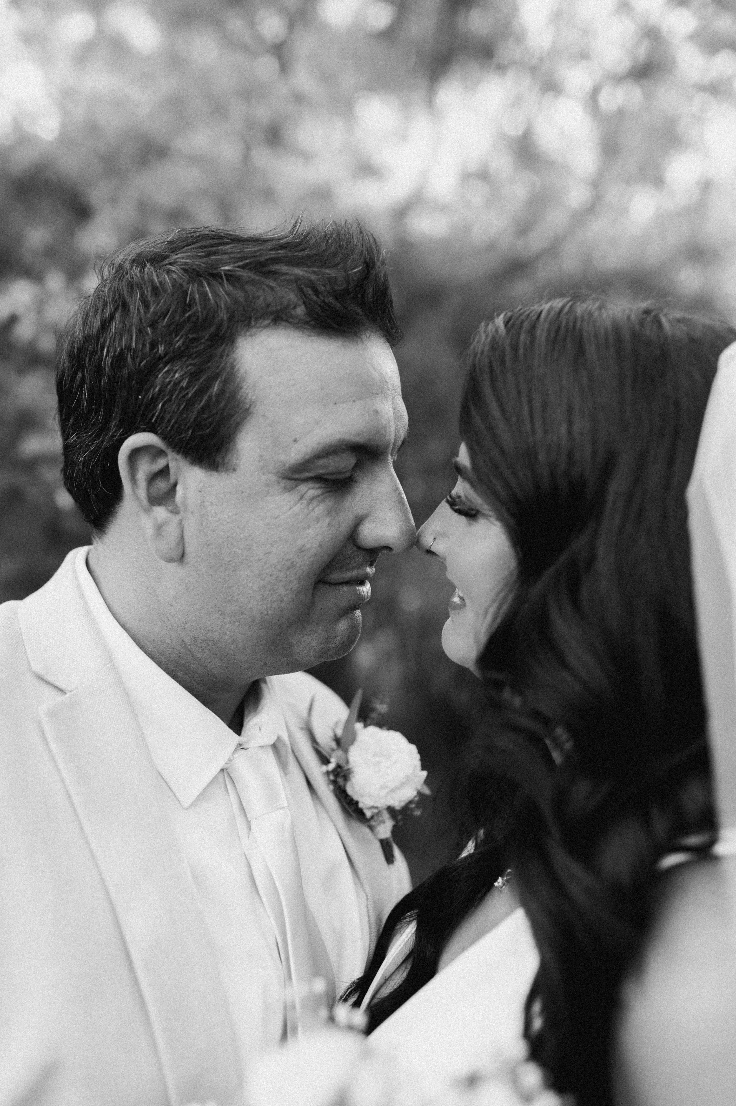 A black and white close-up photo of a groom and bride on their wedding day, nose to nose, smiling, outdoors with blurred trees in the background.