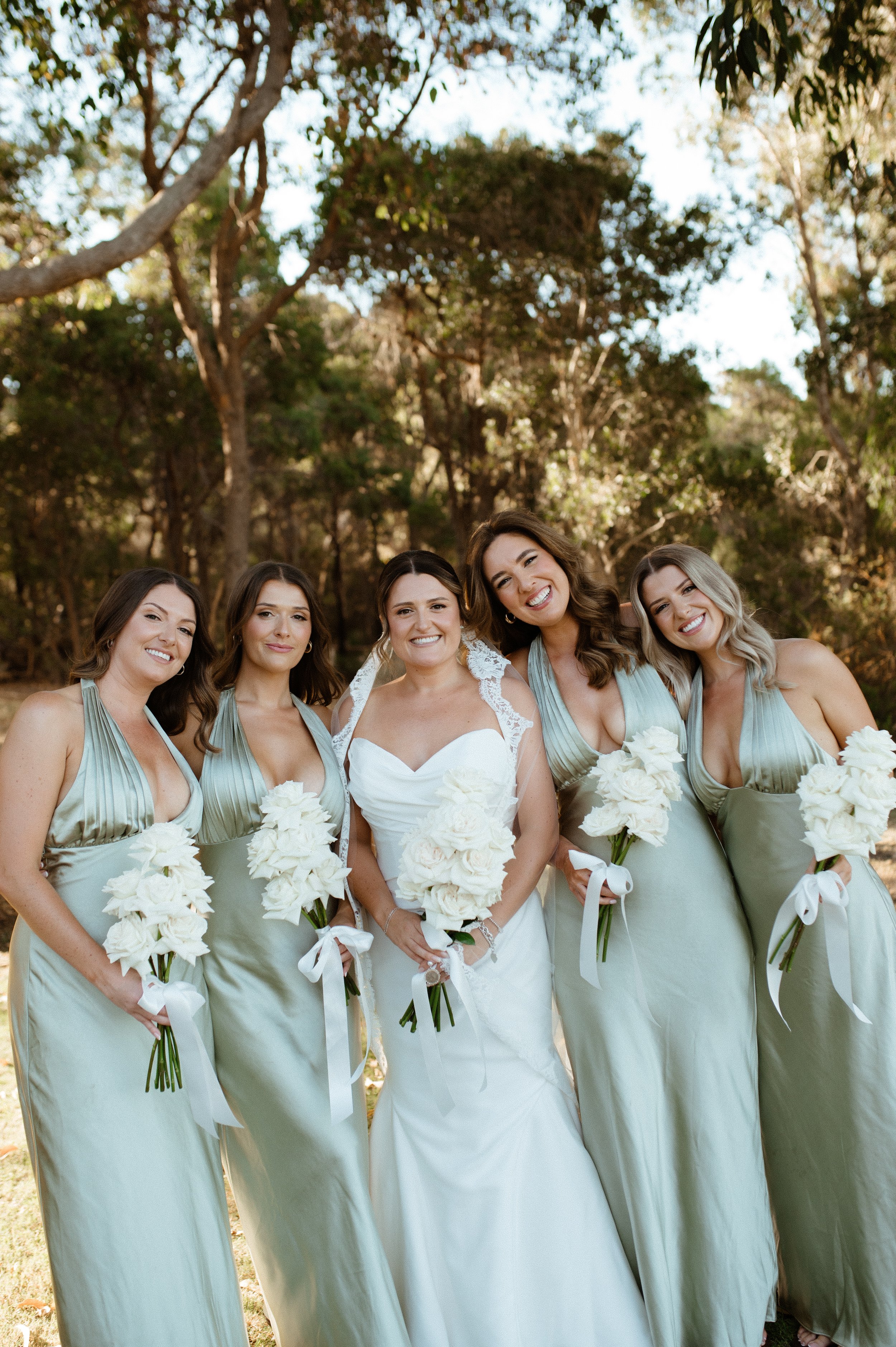 A bride and her four bridesmaids standing outdoors, smiling, each holding a bouquet of white flowers. The bride is in a white wedding dress, and the bridesmaids are in matching light green gowns.