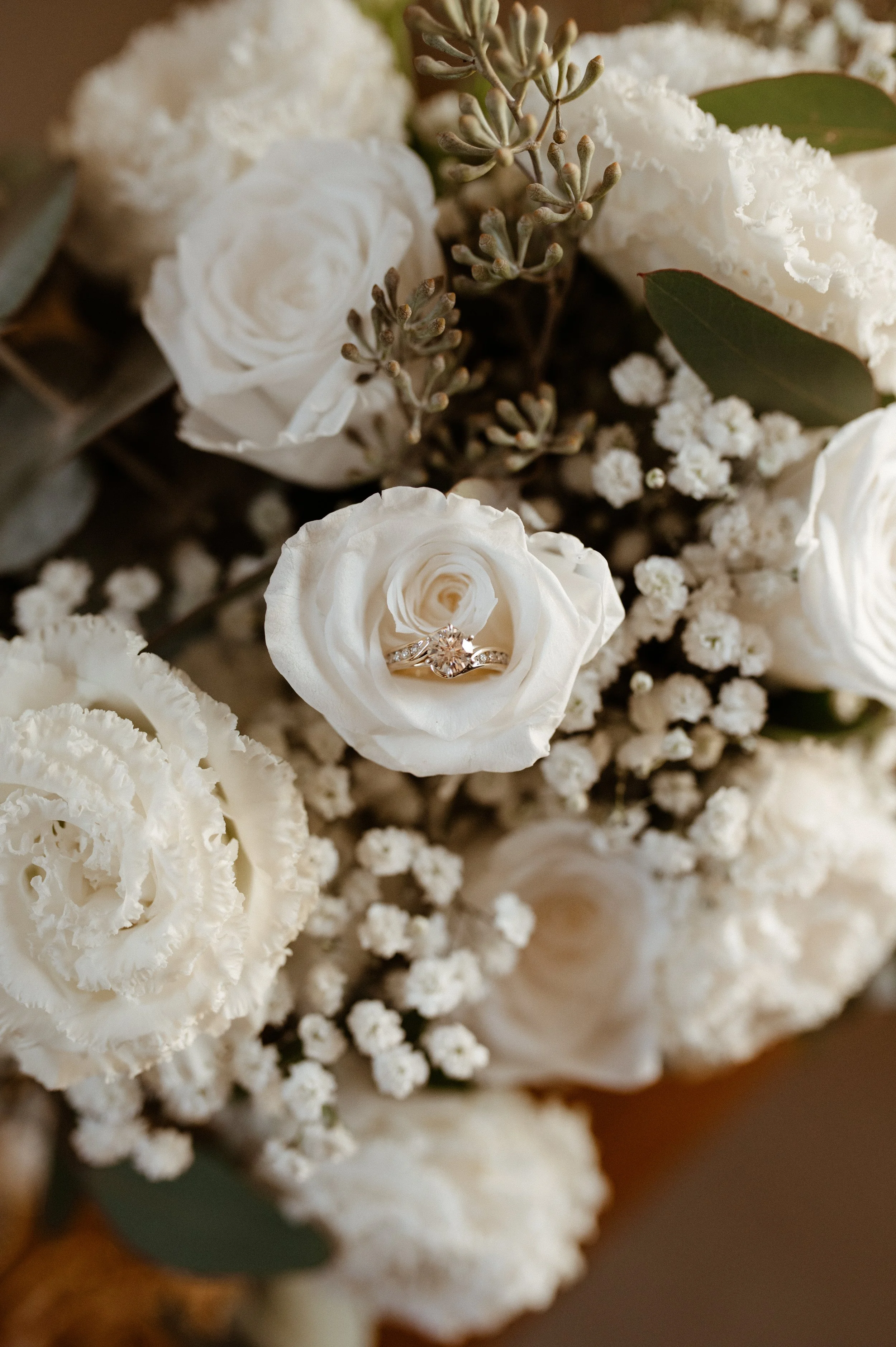 Close-up of a wedding bouquet with white roses, baby's breath, and greenery, featuring a ring resting inside one of the roses.