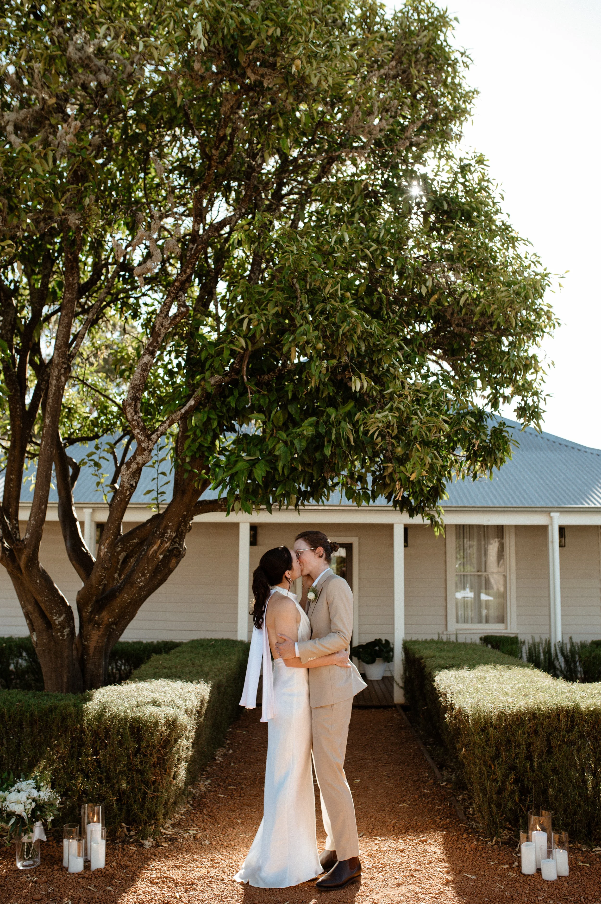Two women wearing wedding dresses sharing a kiss outdoors in front of a house, under a large tree, with candles and lanterns on the ground, during a wedding or celebration.