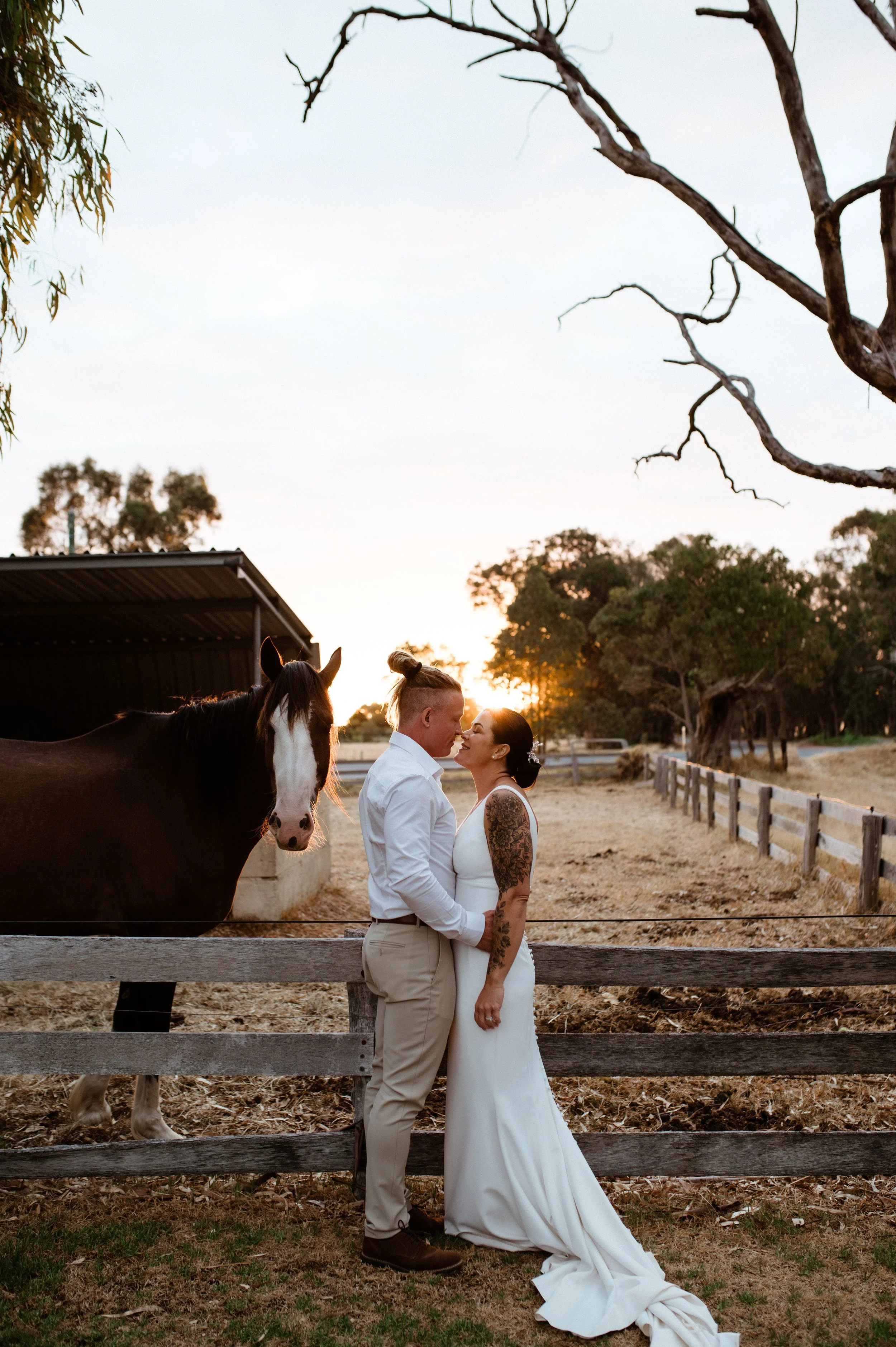 Couple dressed in wedding attire embracing near a horse at sunset on a farm or ranch.