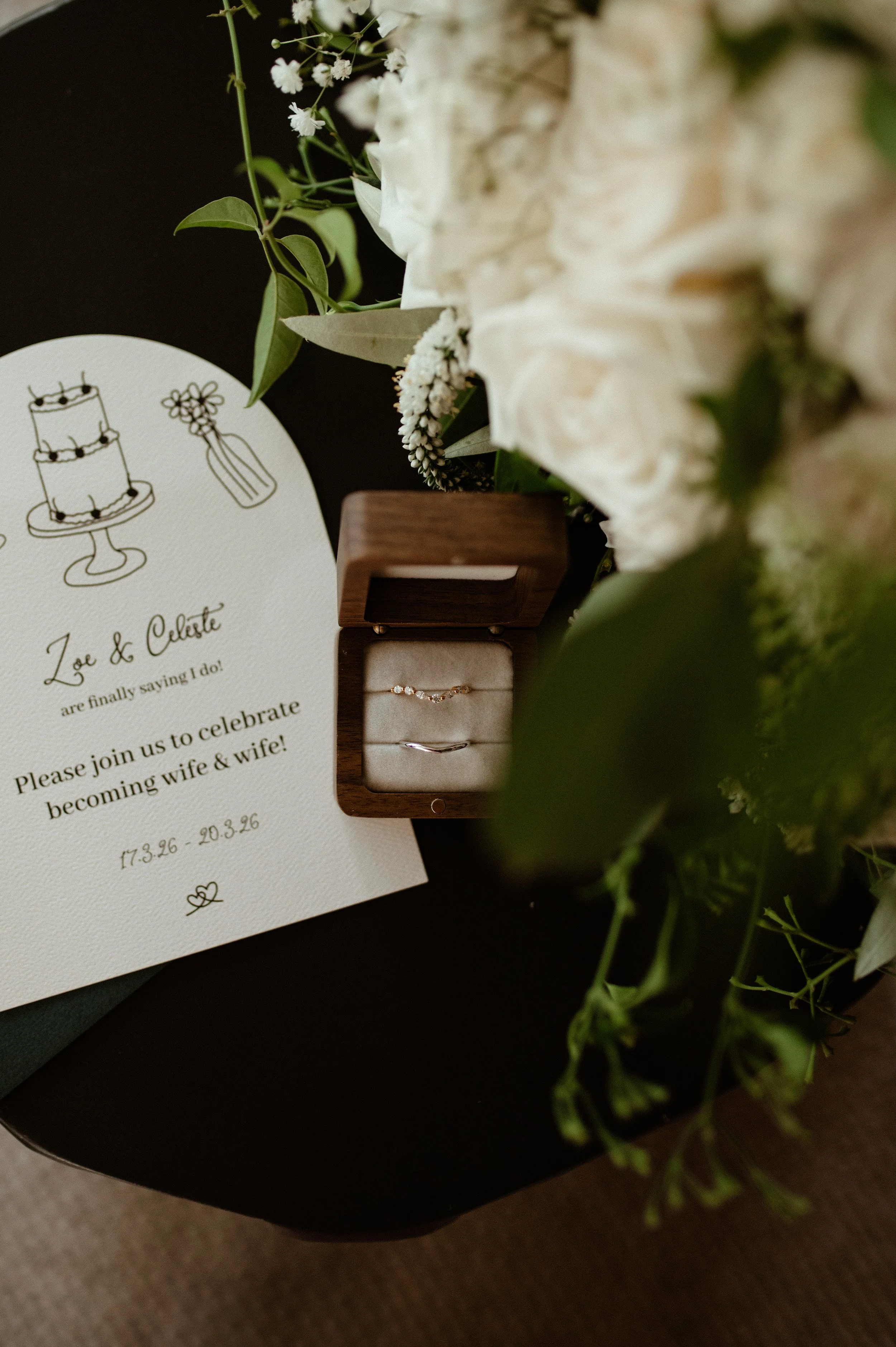 Wedding invitation, engagement rings in a wooden box, and white flowers.