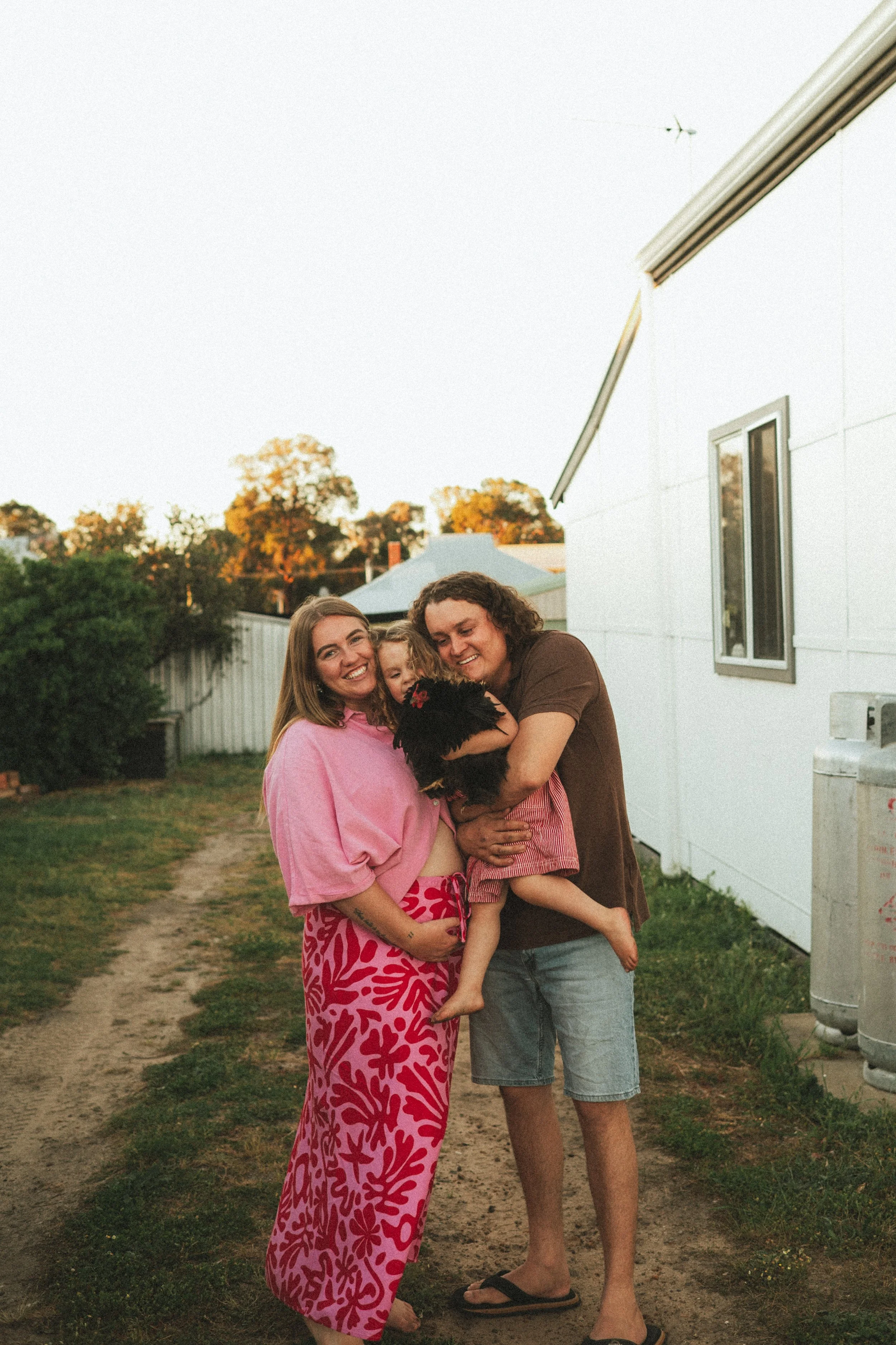 A happy family of three with a dog standing outside on a dirt path at sunset. The woman is wearing a pink blouse and patterned skirt, the man is in a brown T-shirt and shorts, and the young girl is holding a black fluffy dog and is dressed in red and pink pajamas.