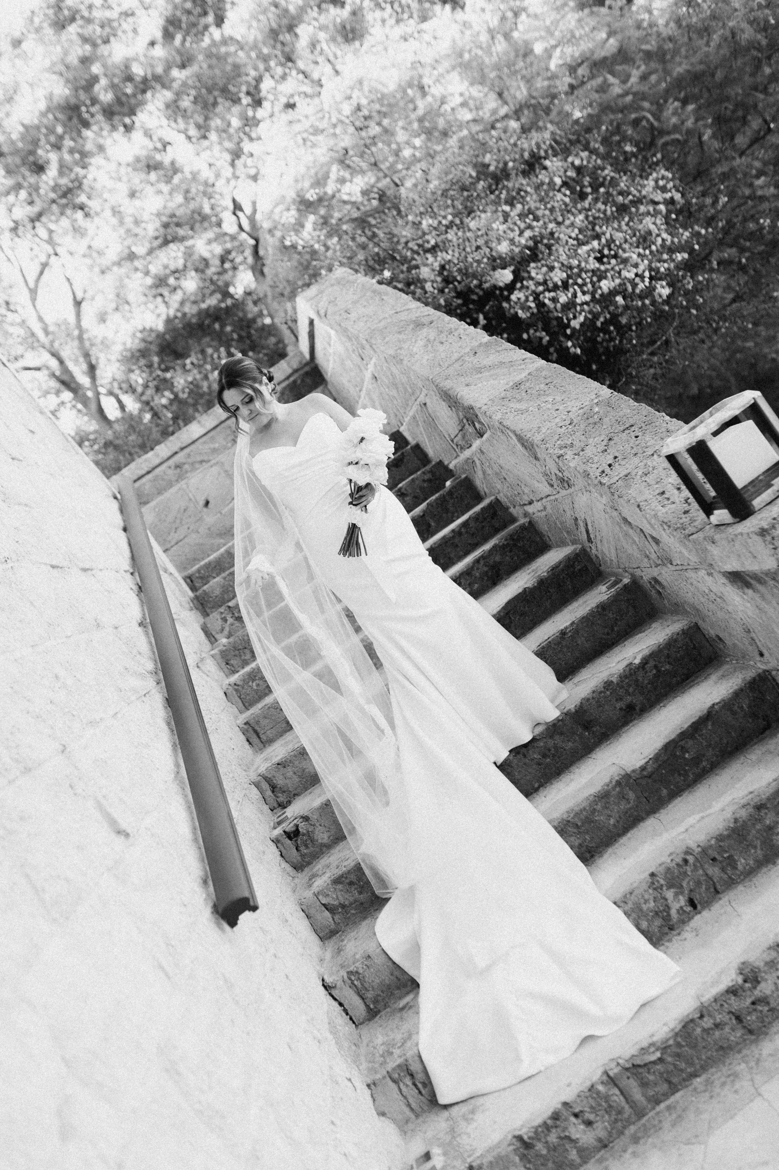 Black and white photo of a bride in a wedding gown standing on outdoor stairs, holding a bouquet, with trees in the background.