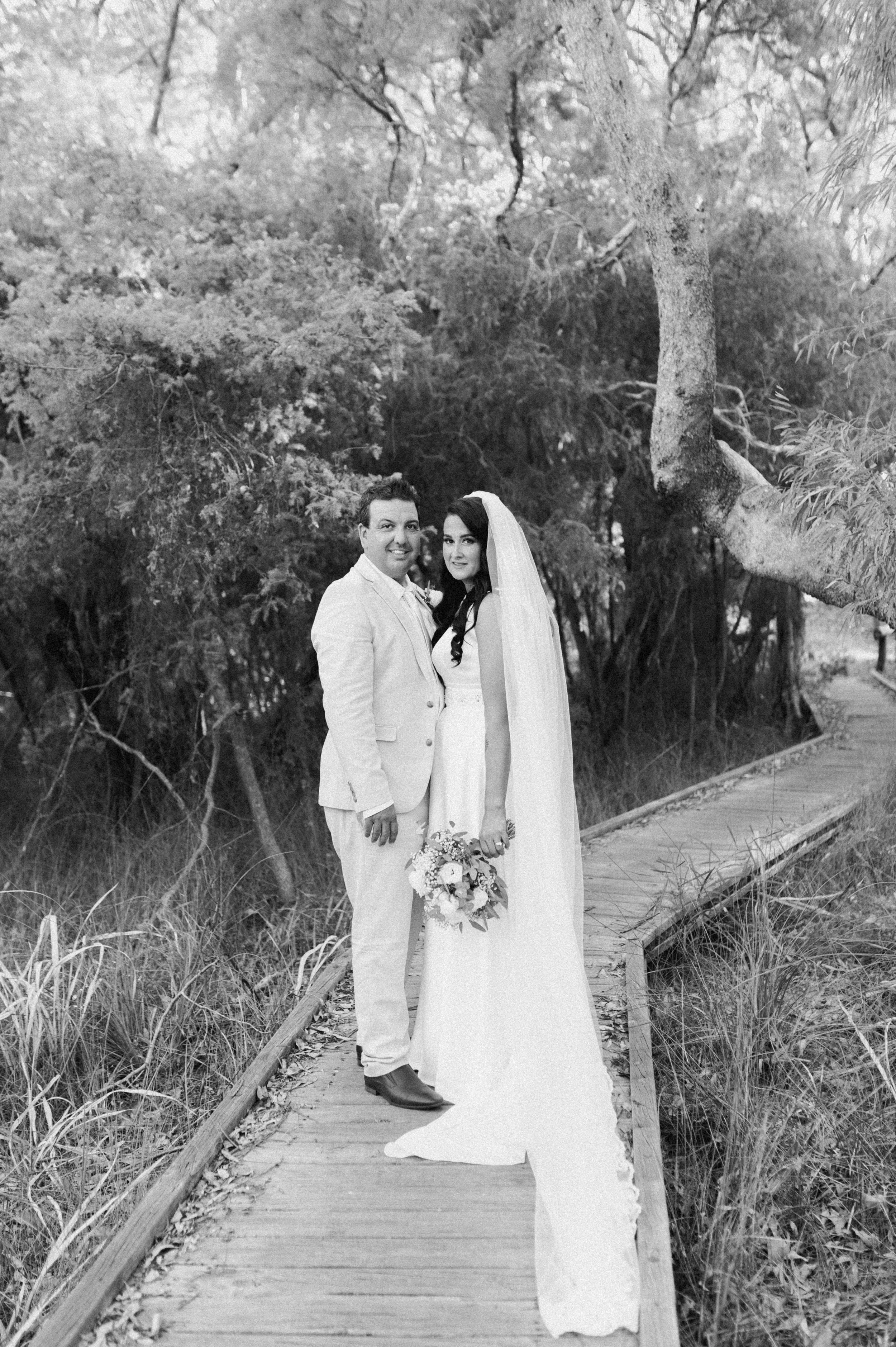 Black and white photograph of a bride and groom standing on a wooden walkway in a natural, wooded setting. The bride is in a wedding dress holding a bouquet, and the groom is in a light-colored suit.