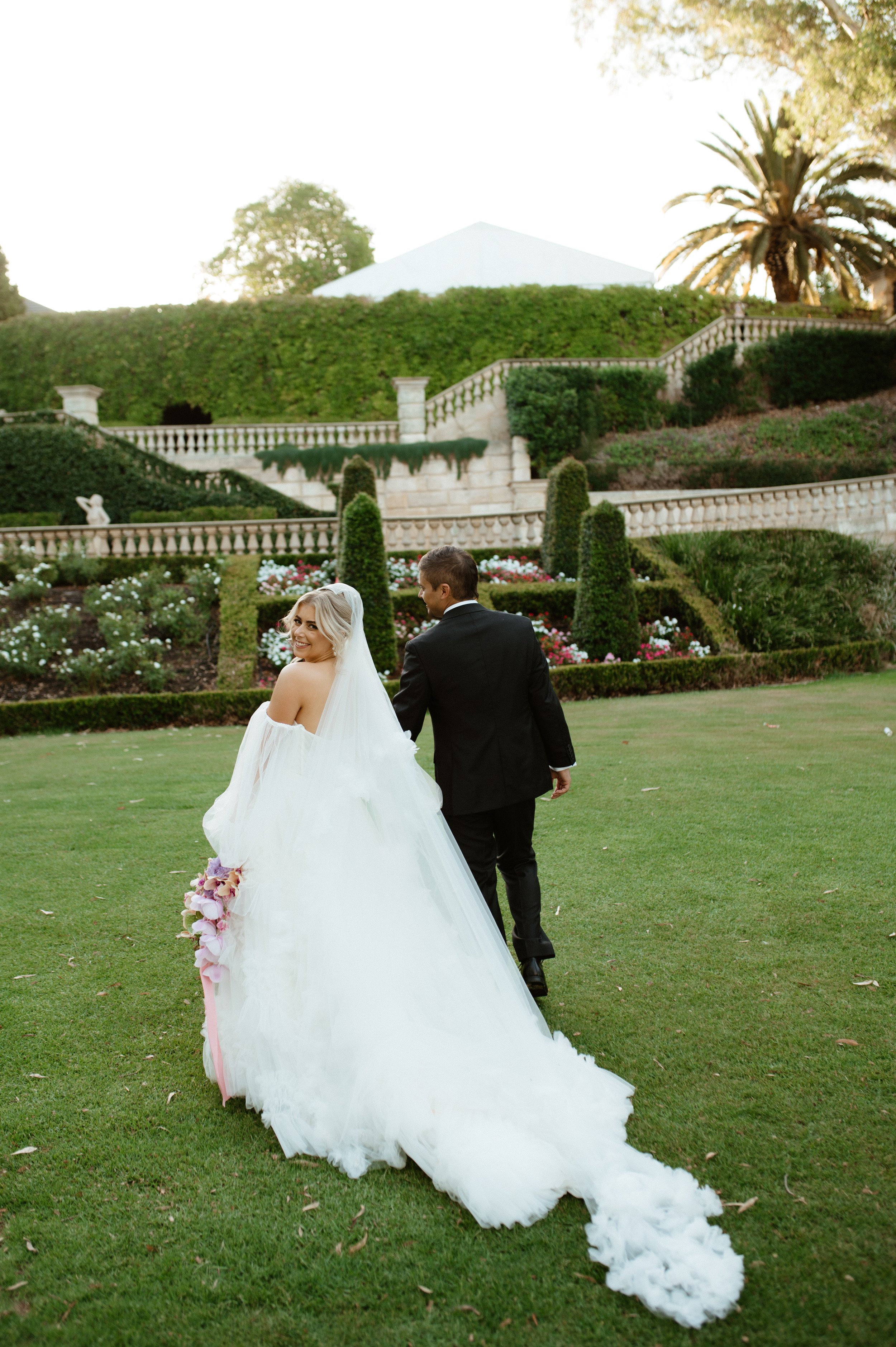 Bride in wedding gown holding bouquet walking on grassy lawn with groom in formal suit, garden with ornamental shrubs and stairs in background.