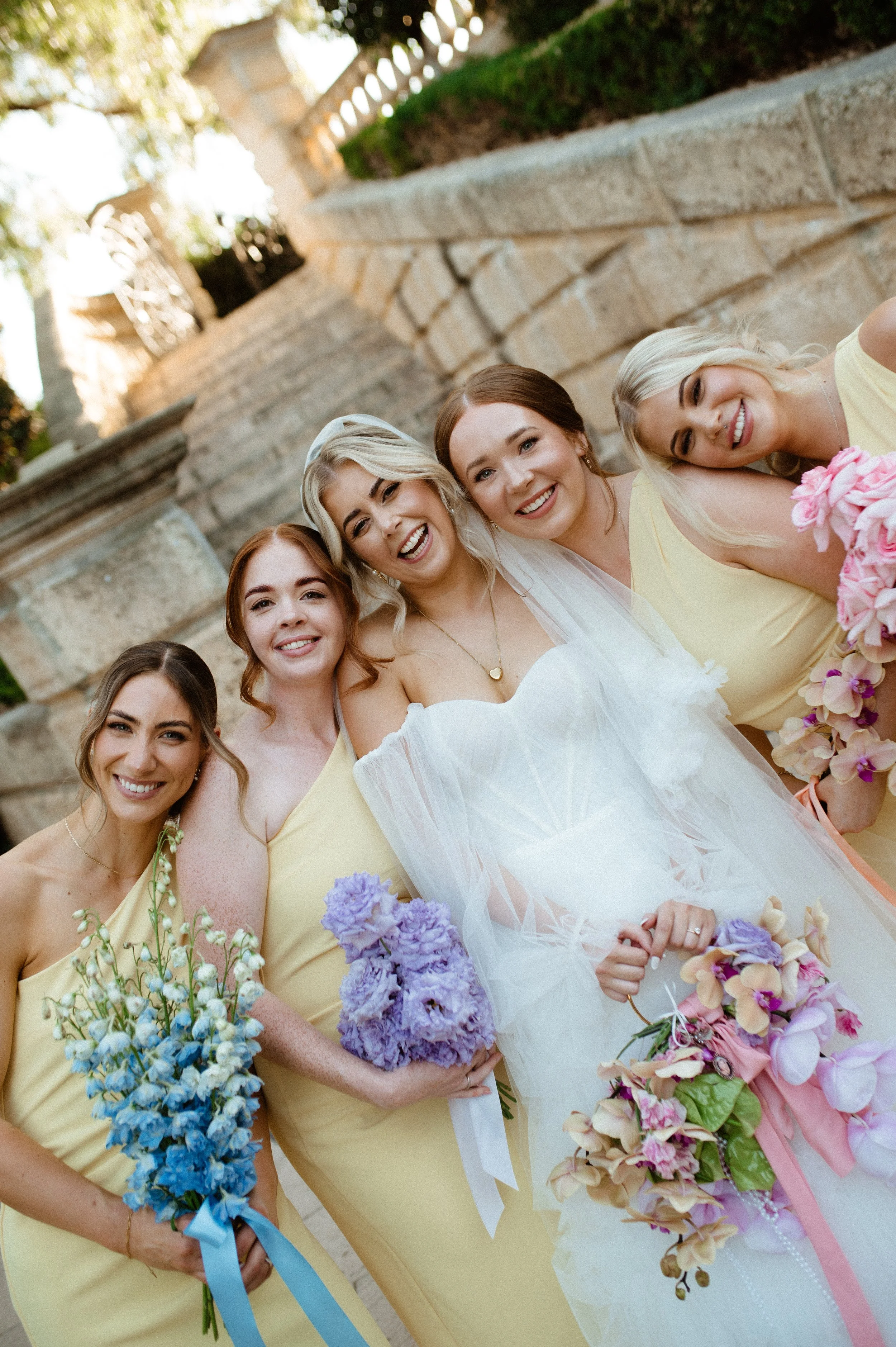 Group of women, including a bride in a white wedding dress and veil, smiling and holding bouquets of flowers, posing together outdoors on stone steps.