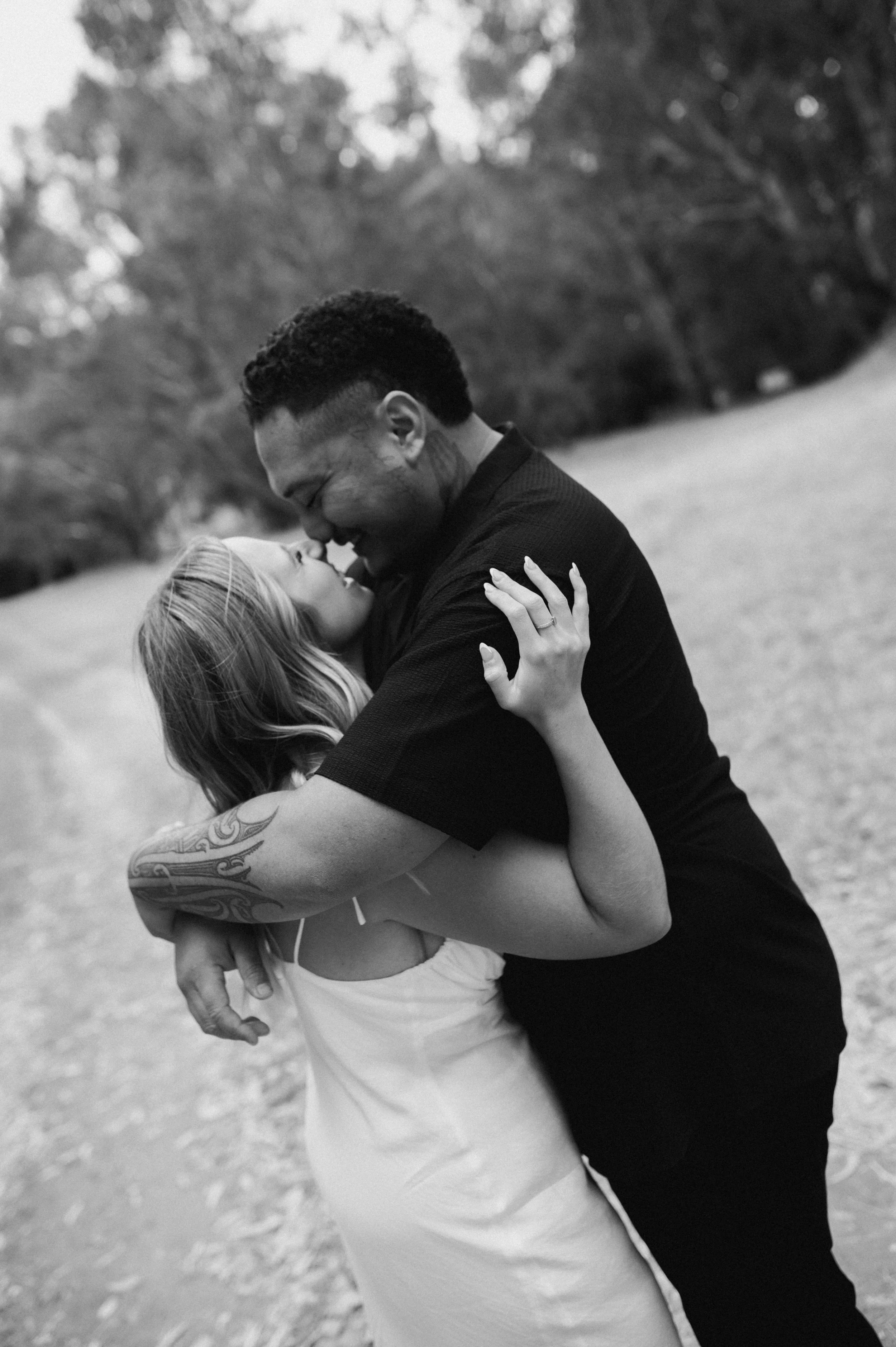 A black and white photo of a couple hugging outdoors, with their faces close together and smiling, in a natural park setting.