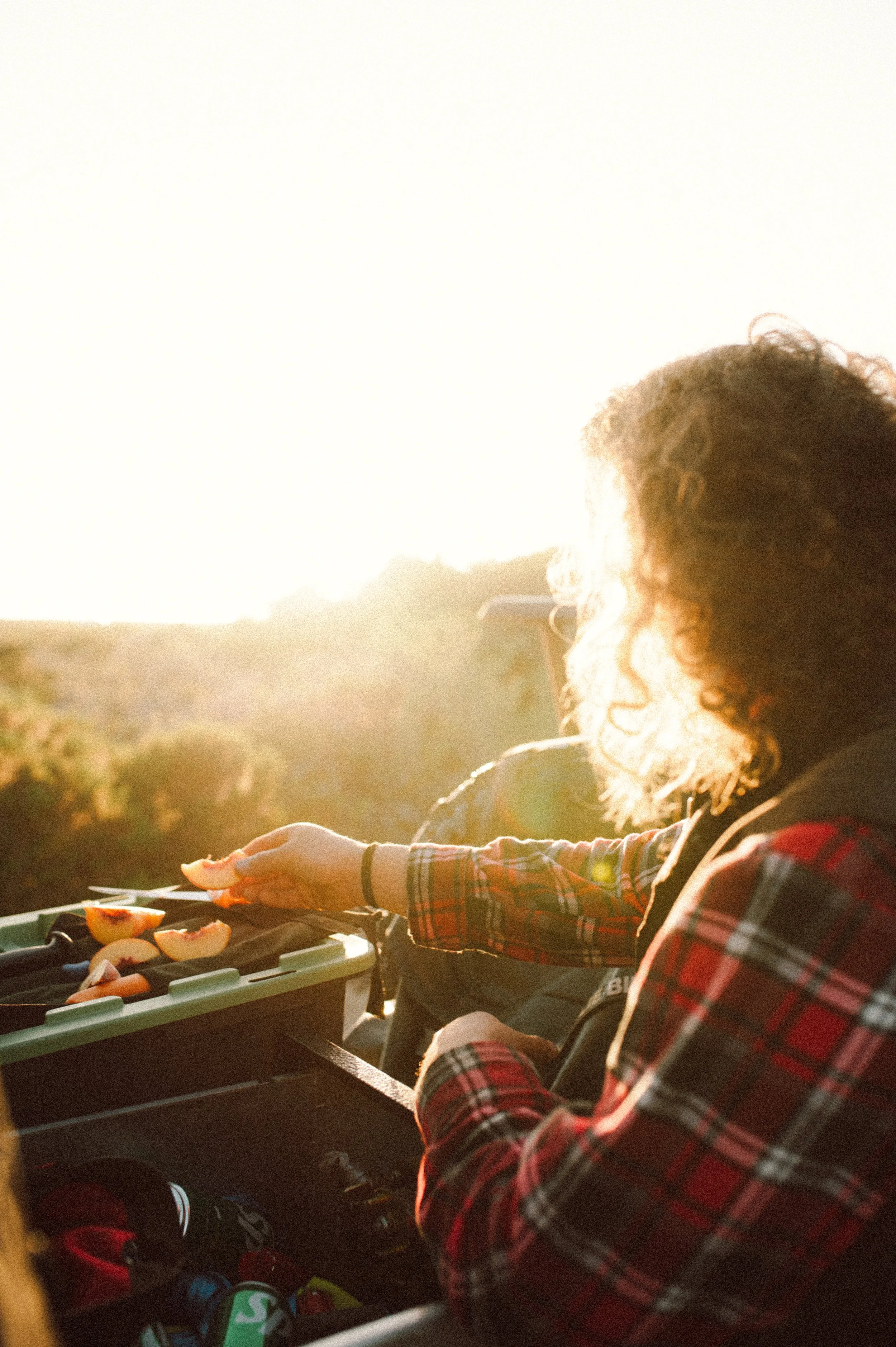 Person with curly hair in a plaid shirt preparing food at sunset or sunrise outdoors.