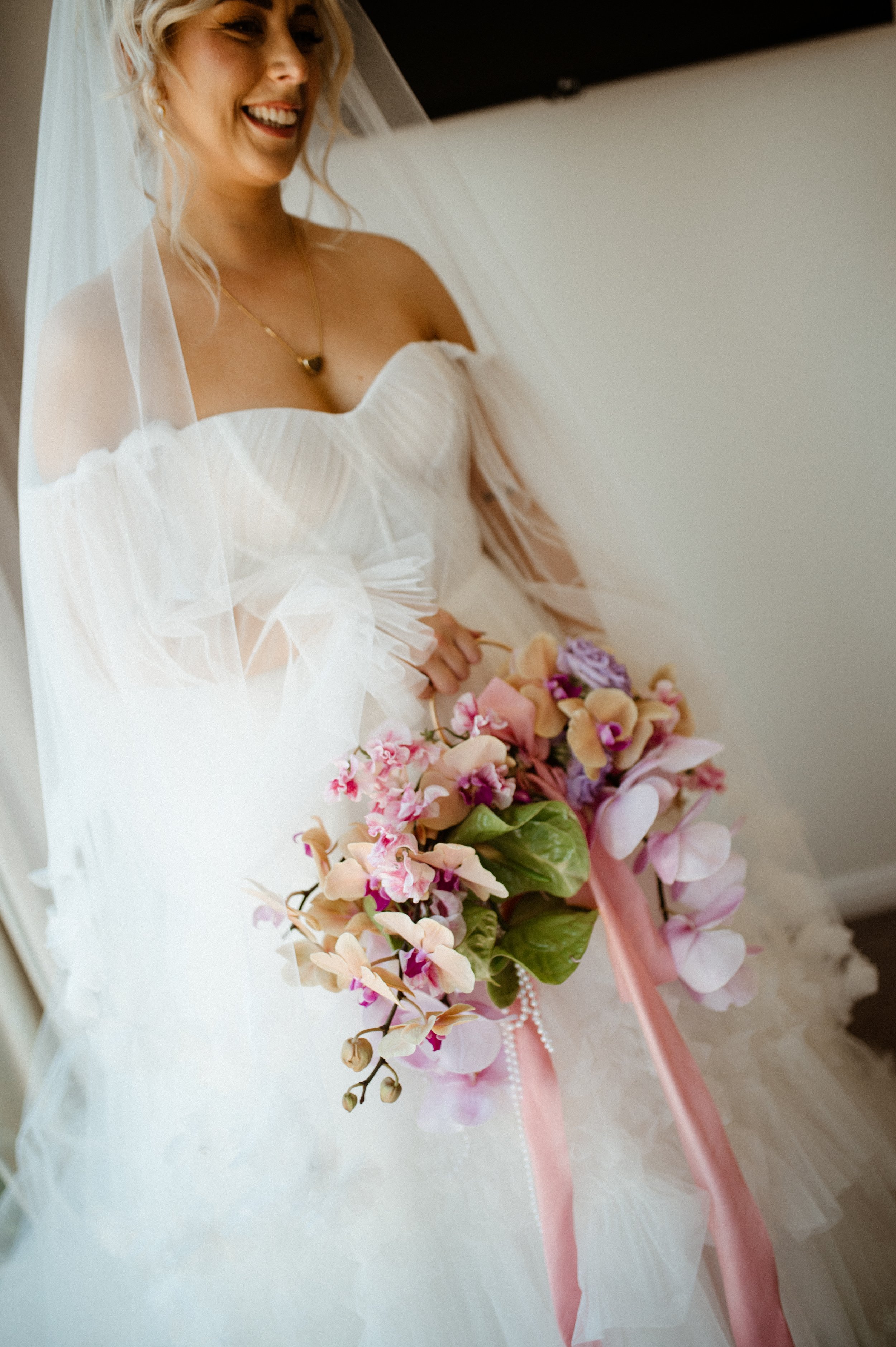 A smiling bride holding a bouquet of pink and purple flowers, wearing a white wedding gown and veil.