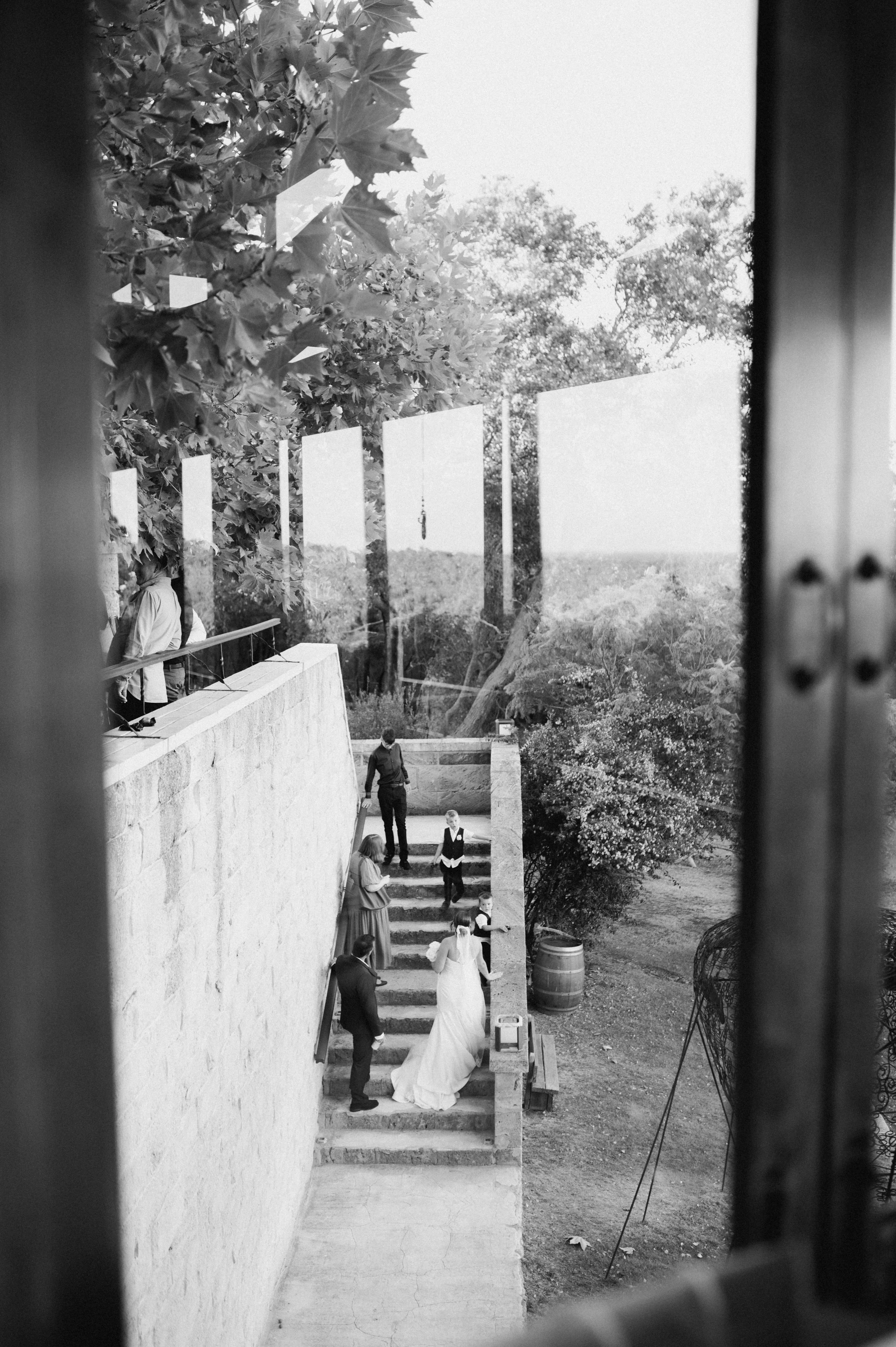 Black and white photo taken through a window showing a woman in a wedding dress and groom walking down stone steps, surrounded by wedding guests, outdoor trees, and greenery.