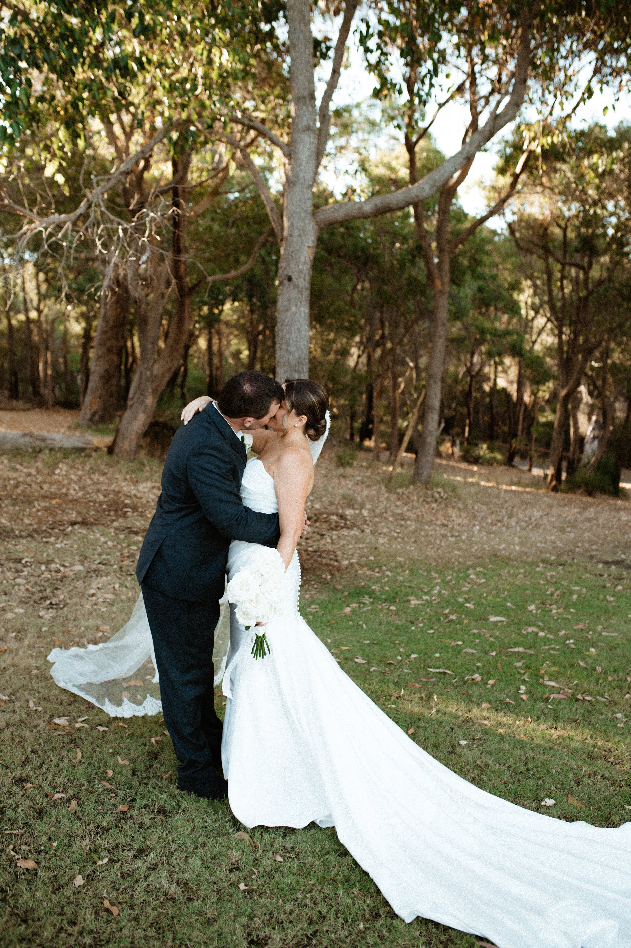 A bride and groom kissing outdoors in a wooded area, with the bride holding a bouquet of white flowers and wearing a white wedding gown with a long train.