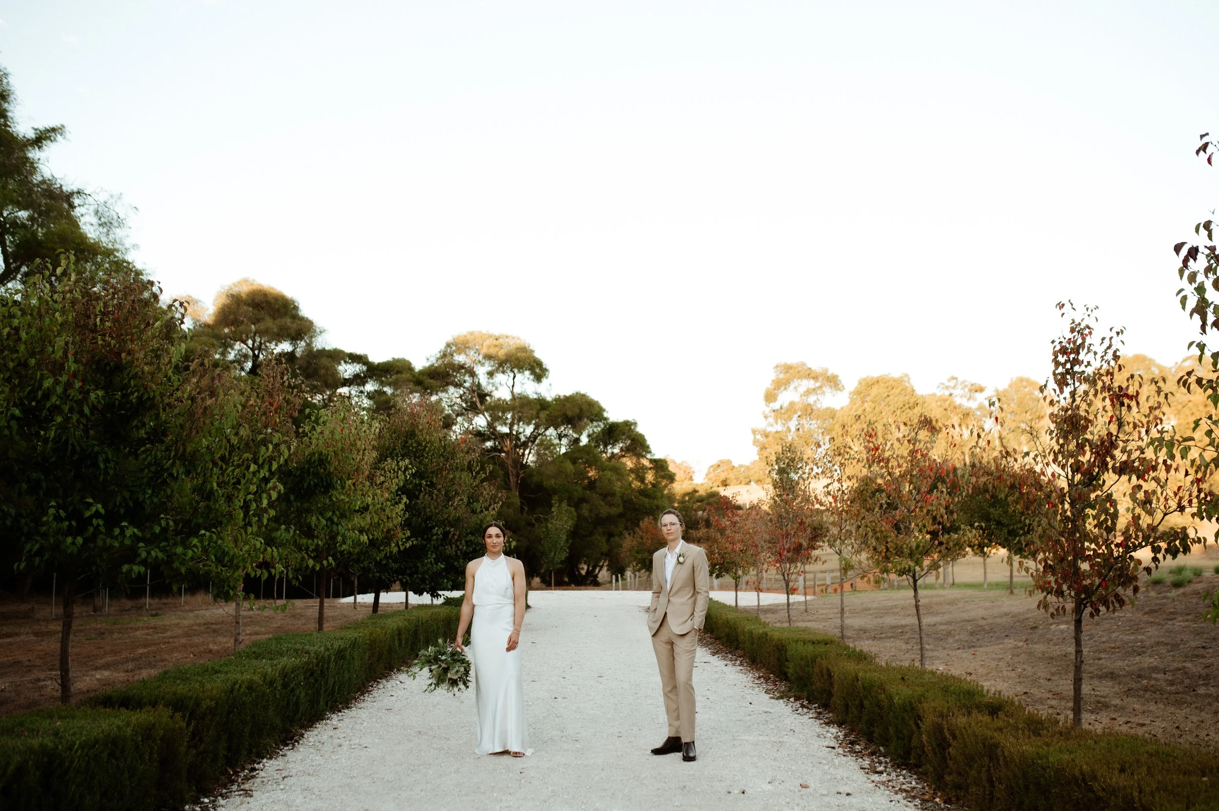 A bride and groom standing on a gravel pathway lined with green bushes and trees, in an outdoor setting during sunset or dusk.