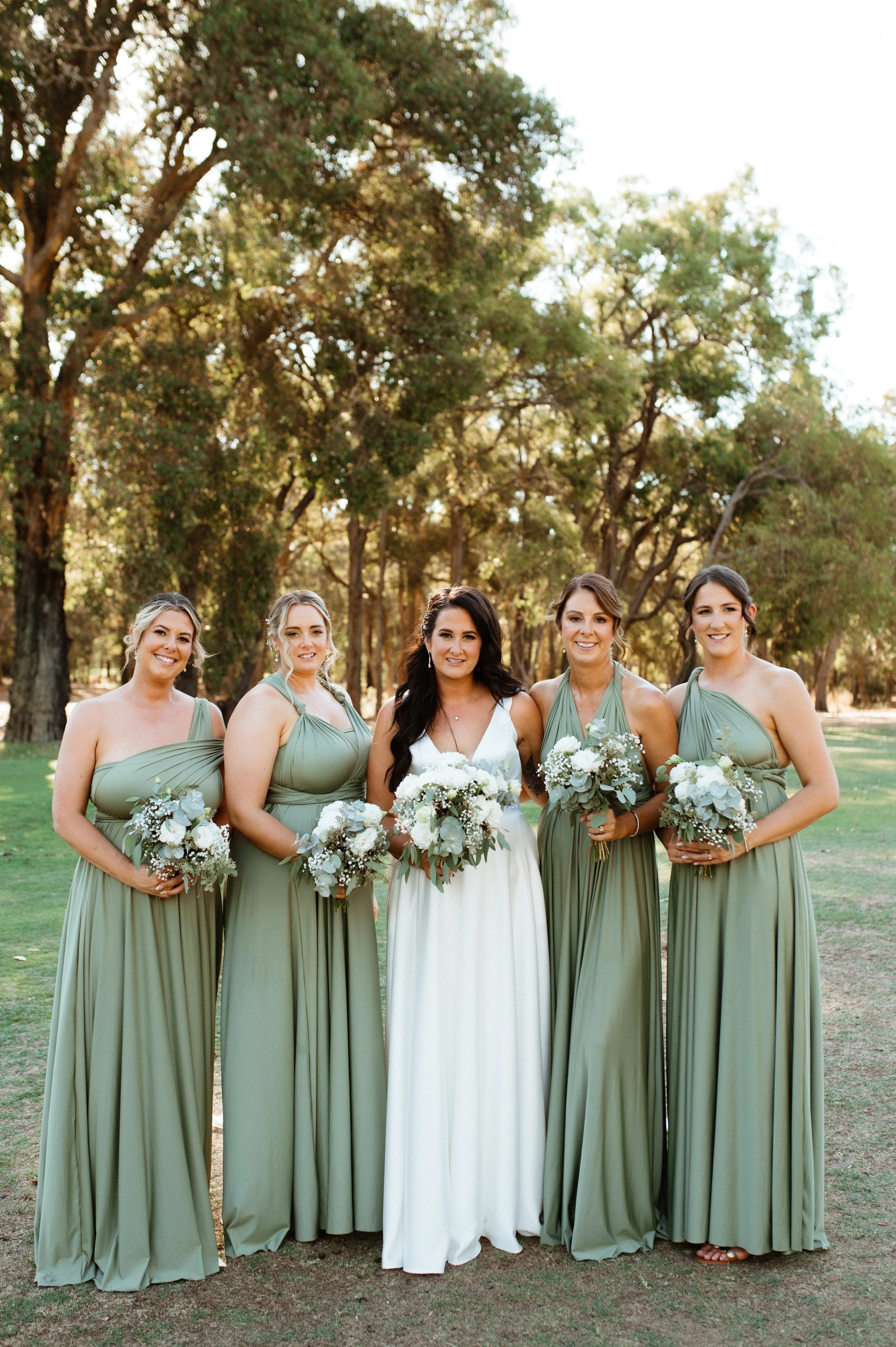 A group of five women, including a bride in a white dress and four bridesmaids in matching sage green dresses, standing outdoors on a grassy area with trees in the background, holding white floral bouquets.