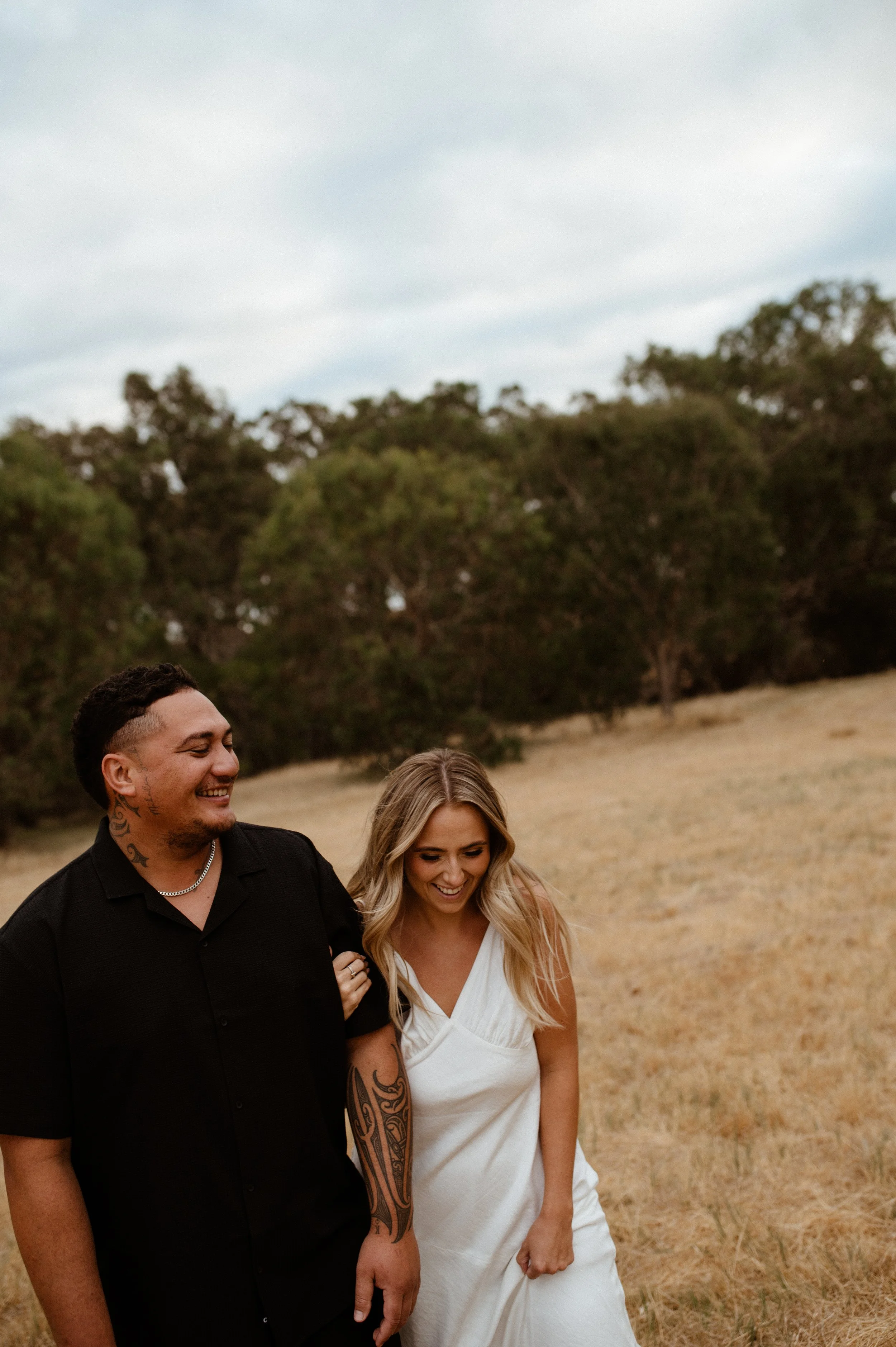 A couple walking and smiling outdoors on a grassy field with trees in the background, overcast sky.