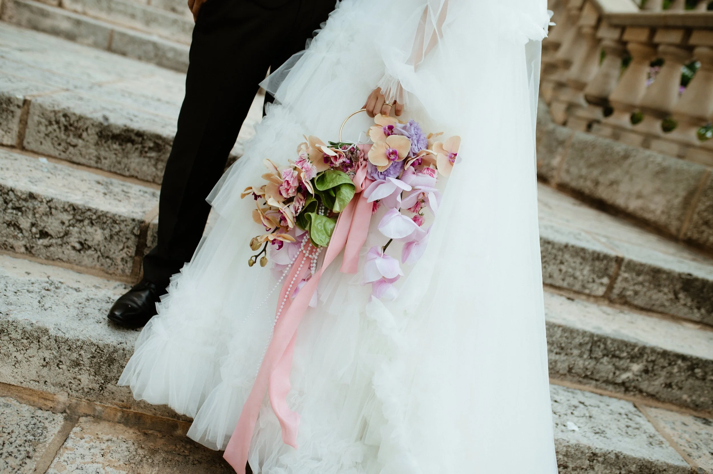 A bride in a white wedding dress holding a bouquet of pink, purple, and peach flowers with pink ribbons, standing on stone steps.