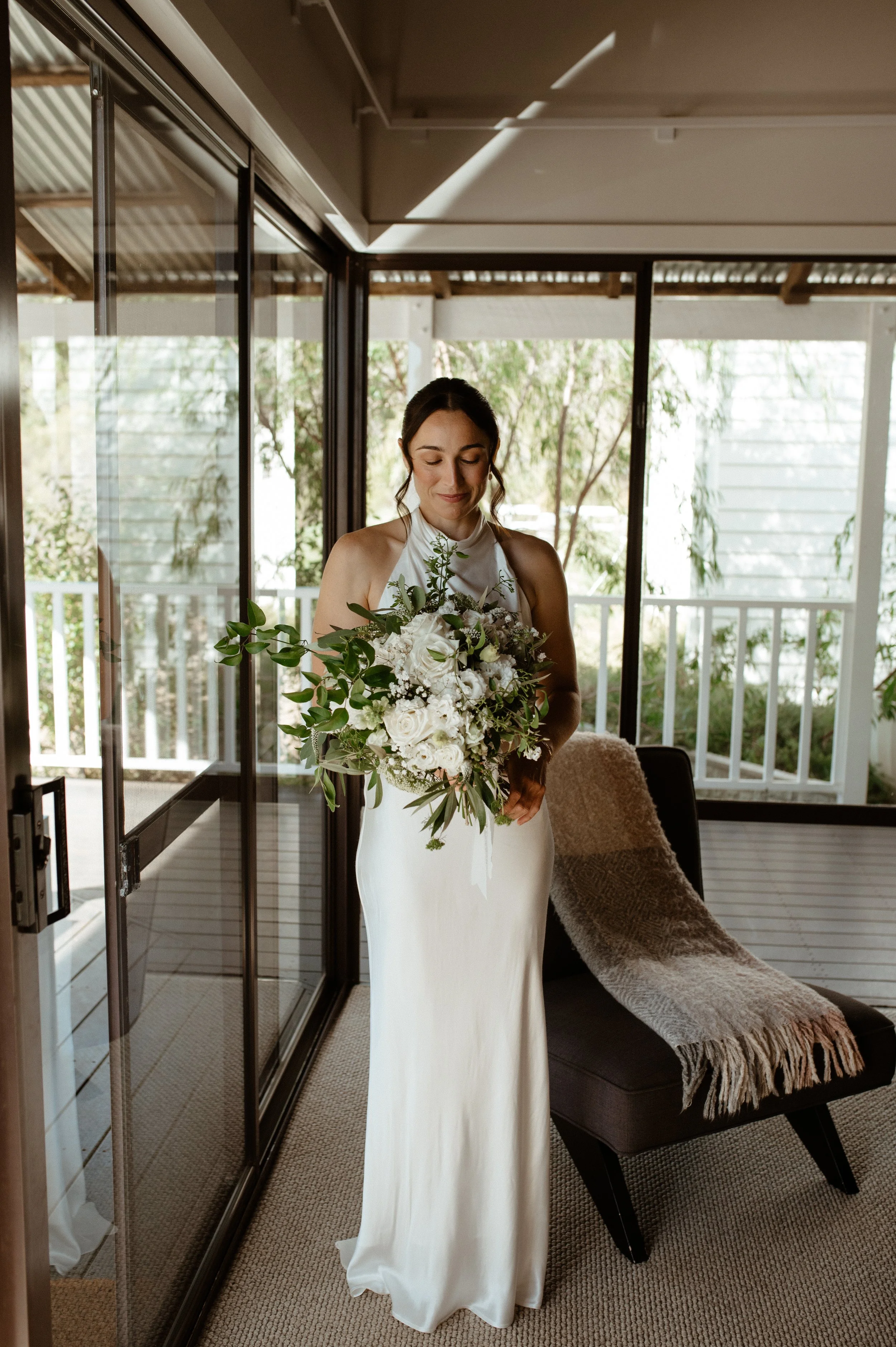 A bride standing indoors, holding a bouquet of white and green flowers, wearing a white satin dress with a halter neckline, with a cozy armchair and a blanket behind her, natural light coming through large glass doors.