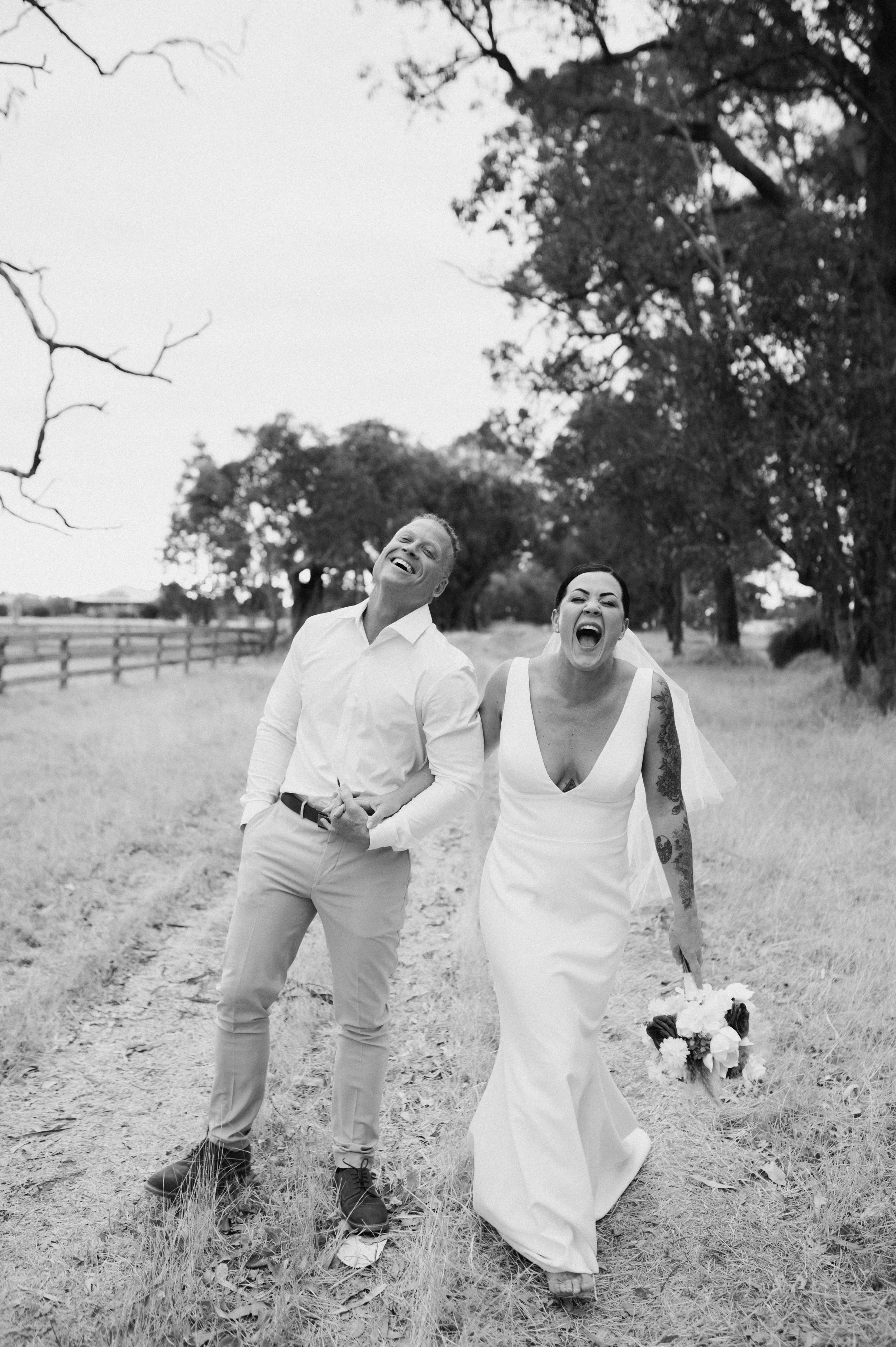 Black and white photo of a bride and groom walking outdoors, laughing and smiling, with trees and a fence in the background.
