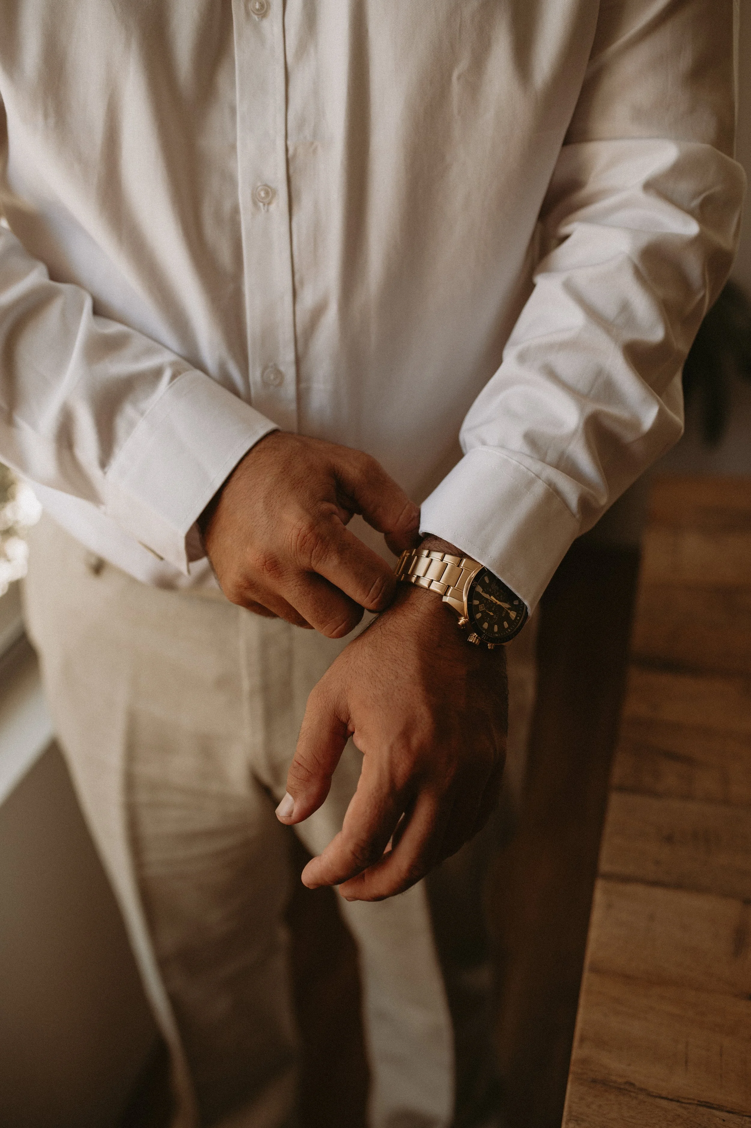 Close-up of a man wearing a white dress shirt and a gold wristwatch, adjusting the watch on his left wrist, standing near a wooden table.