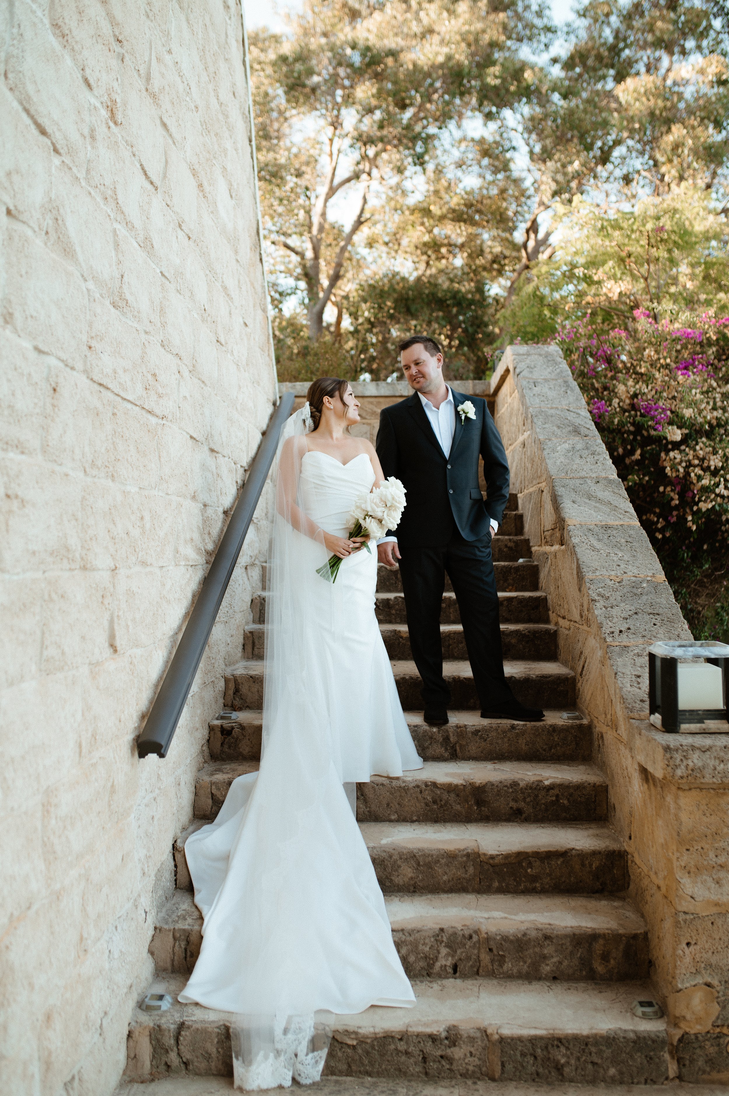 A bride and groom standing on stone steps outdoors, with the bride holding a bouquet of white flowers and wearing a white wedding dress, and the groom in a dark suit with a white shirt and boutonniere, during a wedding photoshoot.