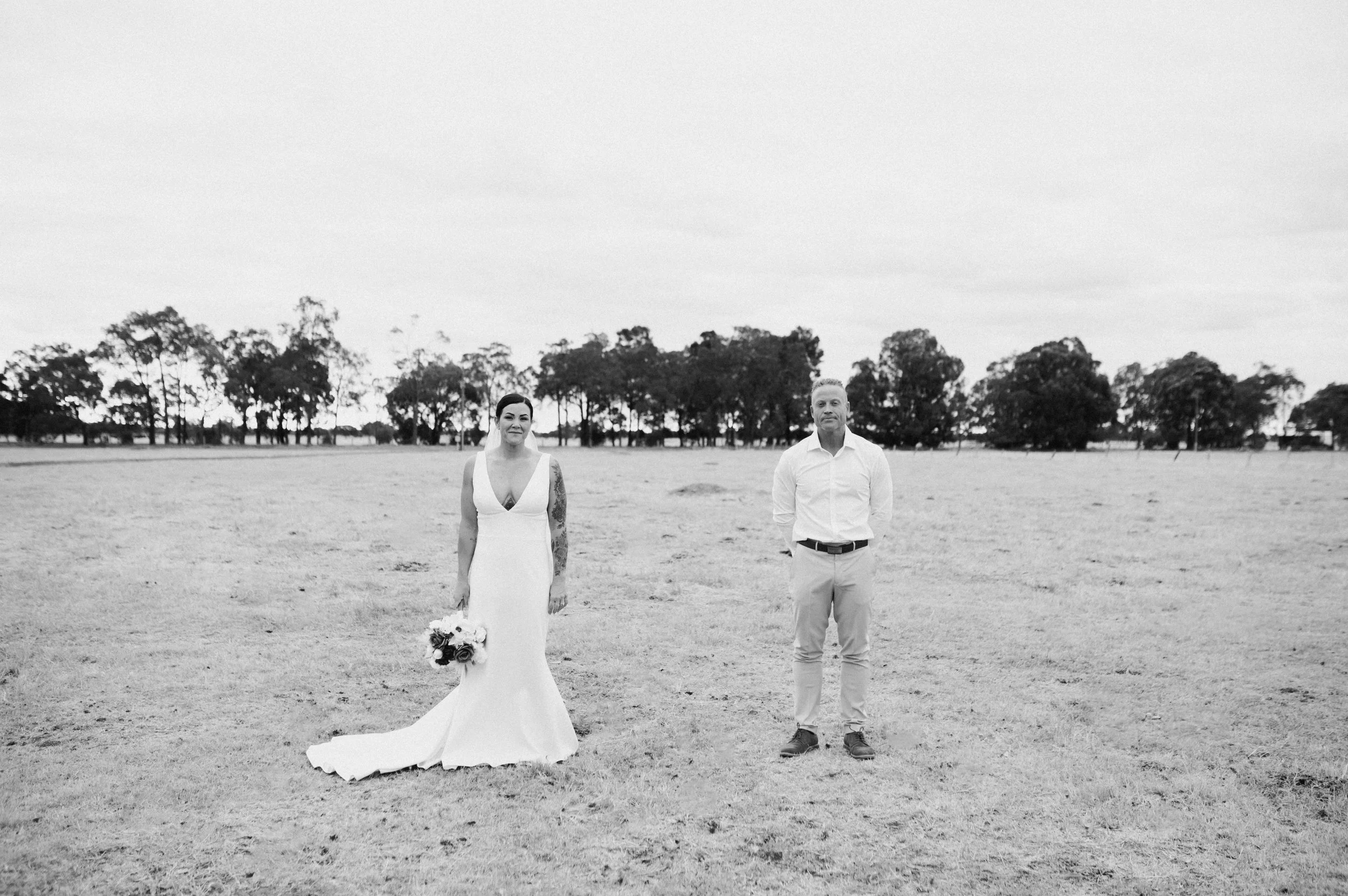 A black and white photo of a bride in a white wedding dress holding a bouquet standing apart from a groom in a white shirt and light-colored pants in an open field with trees in the background.