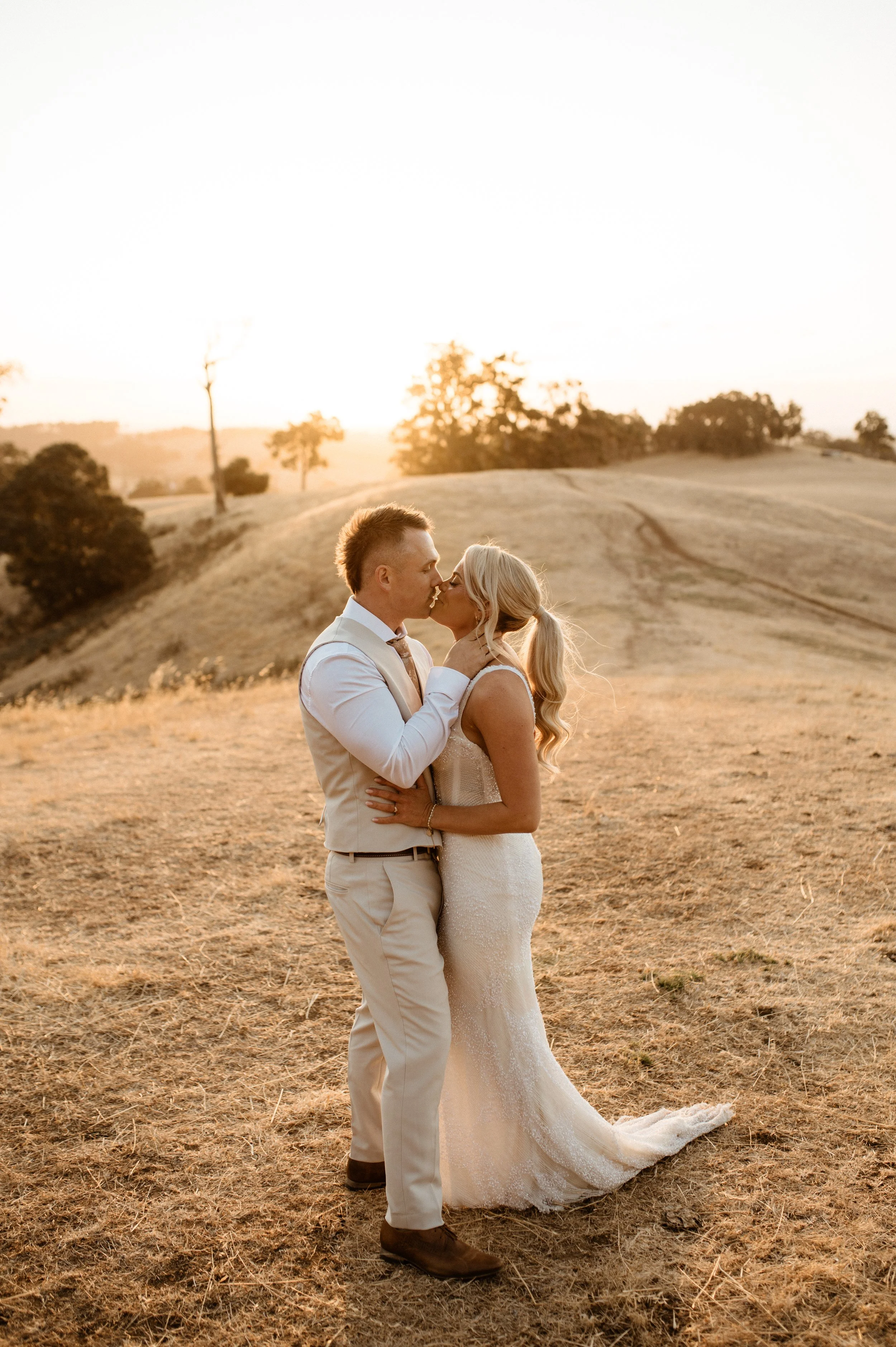 A newlywed couple sharing a kiss outdoors during sunset on a grassy hill.