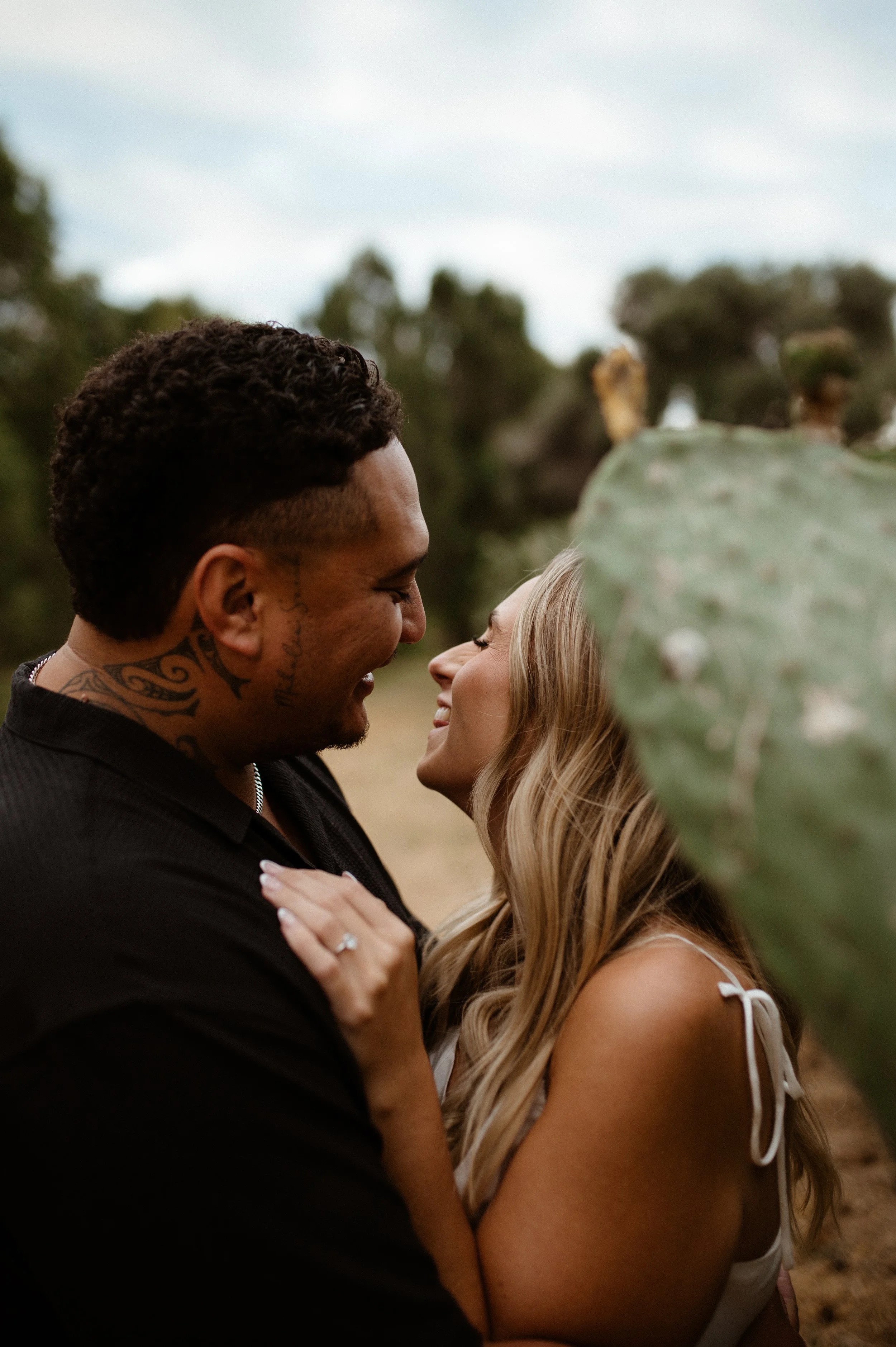 A loving couple standing close, smiling and looking into each other's eyes outdoors on a cloudy day, with blurred trees in the background.