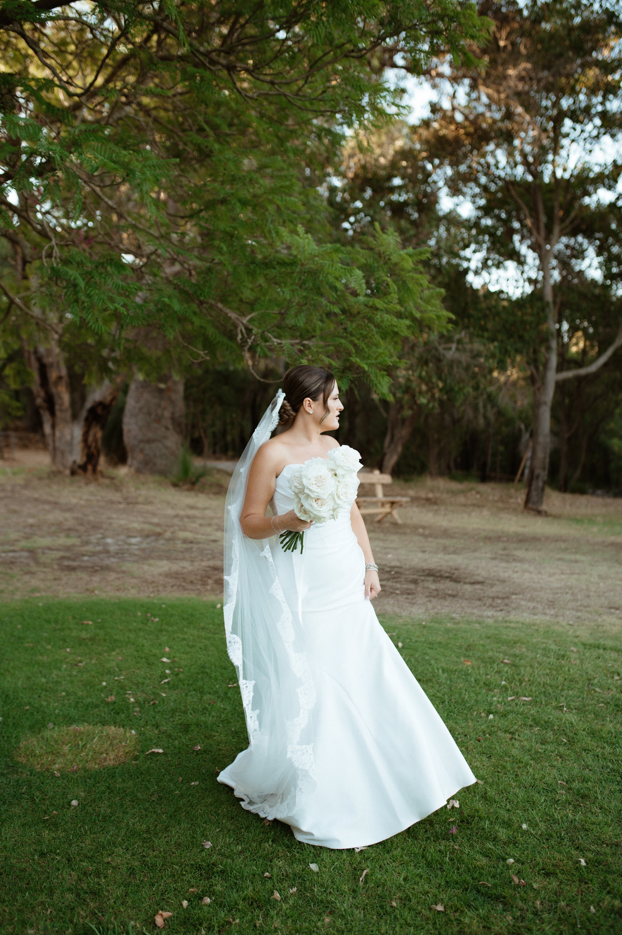 A bride in a white wedding gown and veil holds a bouquet of white roses in a park with green grass and trees.
