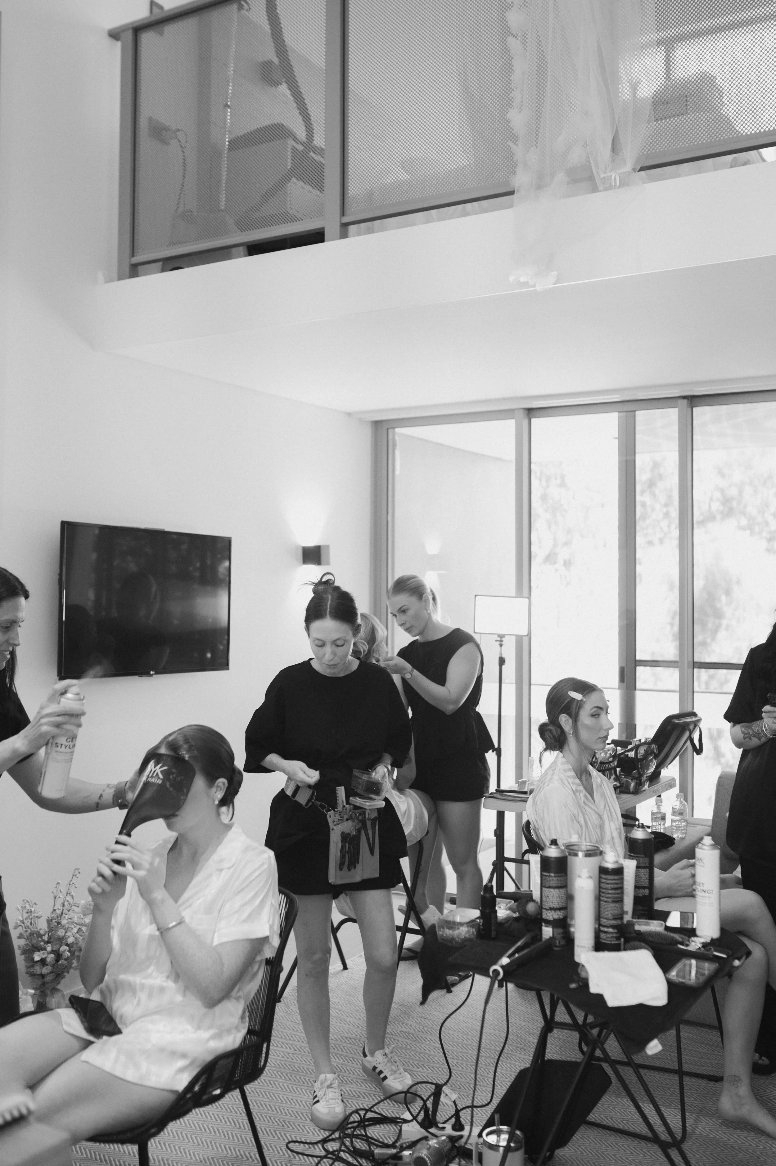 Women preparing models for a photoshoot in a bright dressing room with large windows, makeup and hair styling tools, and a television on the wall.