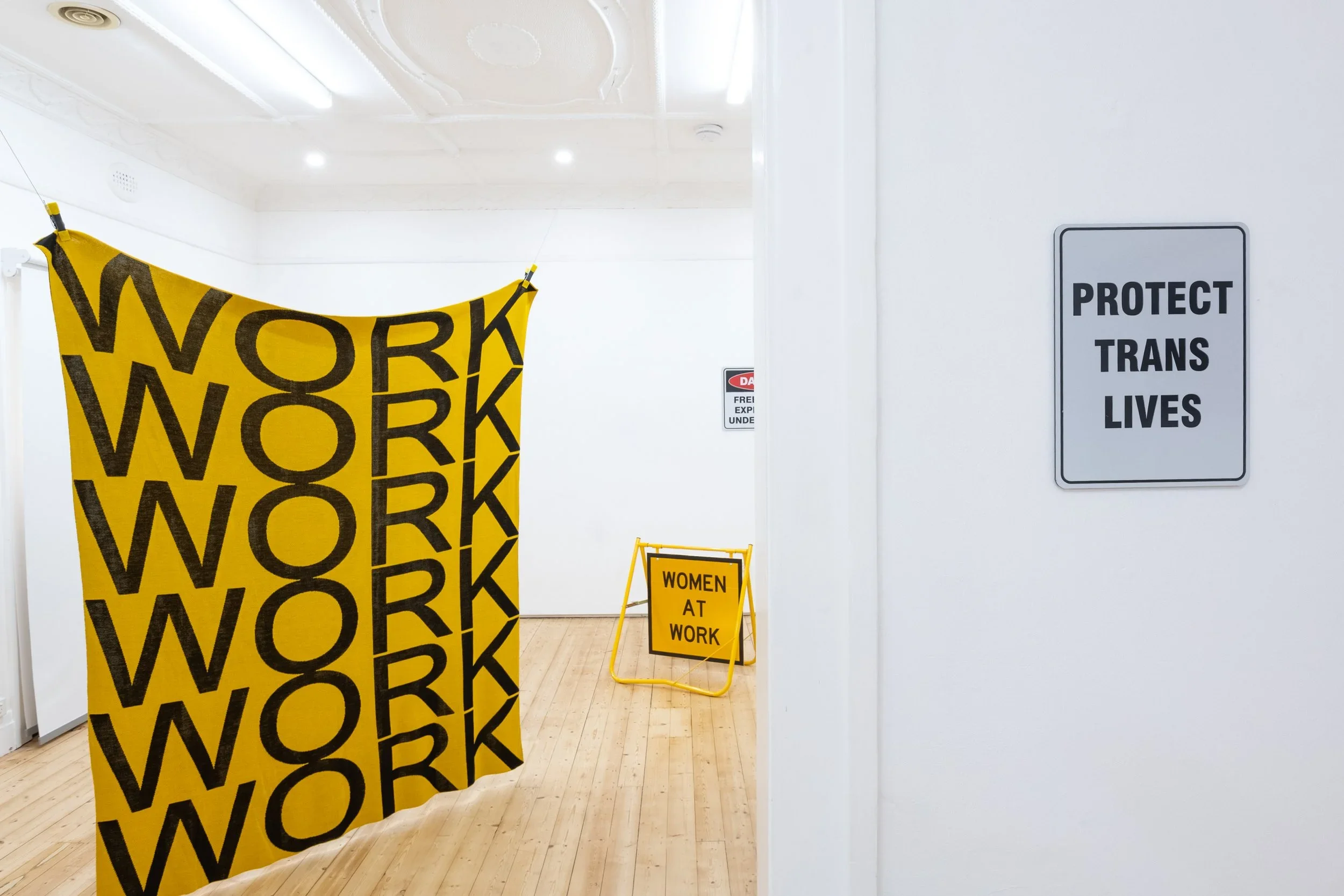 A large knitted banner hangs from the ceiling with the word 'WORK' repeated. A sign hangs to the right reading 'Protect Trans Lives'.